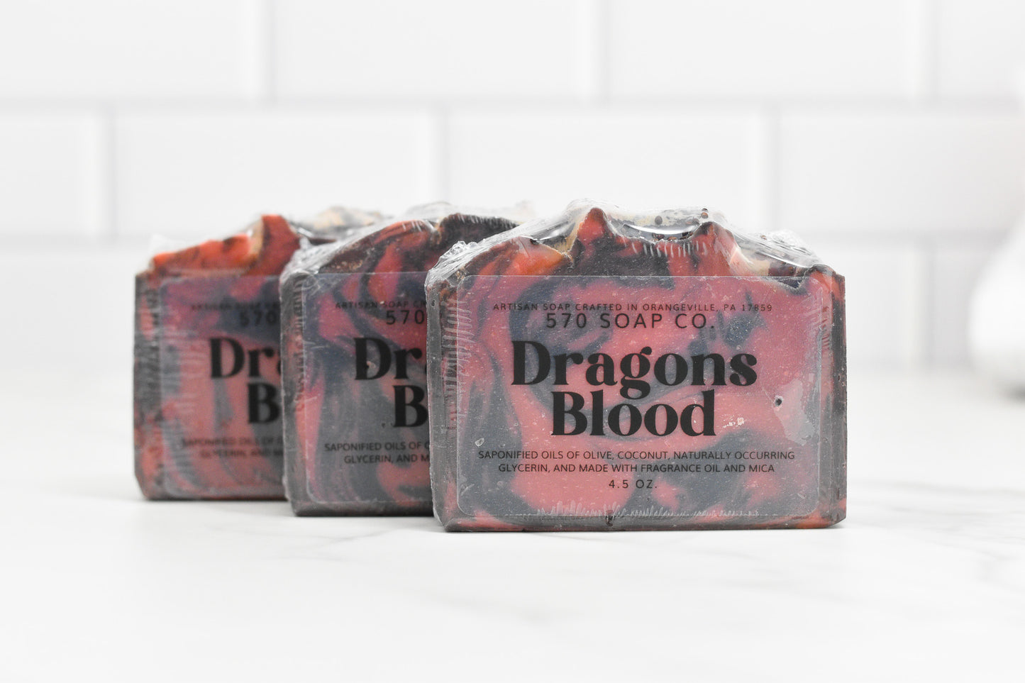 Displayed against a white tile background are three bars of 570 Soap Co.'s Dragons Blood Bar Soap, each infused with an exotic fragrance. The soaps feature a blend of red and black hues, wrapped in clear packaging, and are handcrafted to a quality 4.5 oz size with labeled packaging.