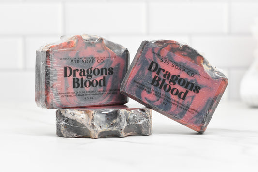 Three bars of Dragons Blood Bar Soap from 570 Soap Co. are displayed, with two wrapped and one unwrapped to reveal a marbled red and black design that exudes an exotic fragrance. They sit on a white marble surface against a tiled backdrop, highlighting their handcrafted quality.