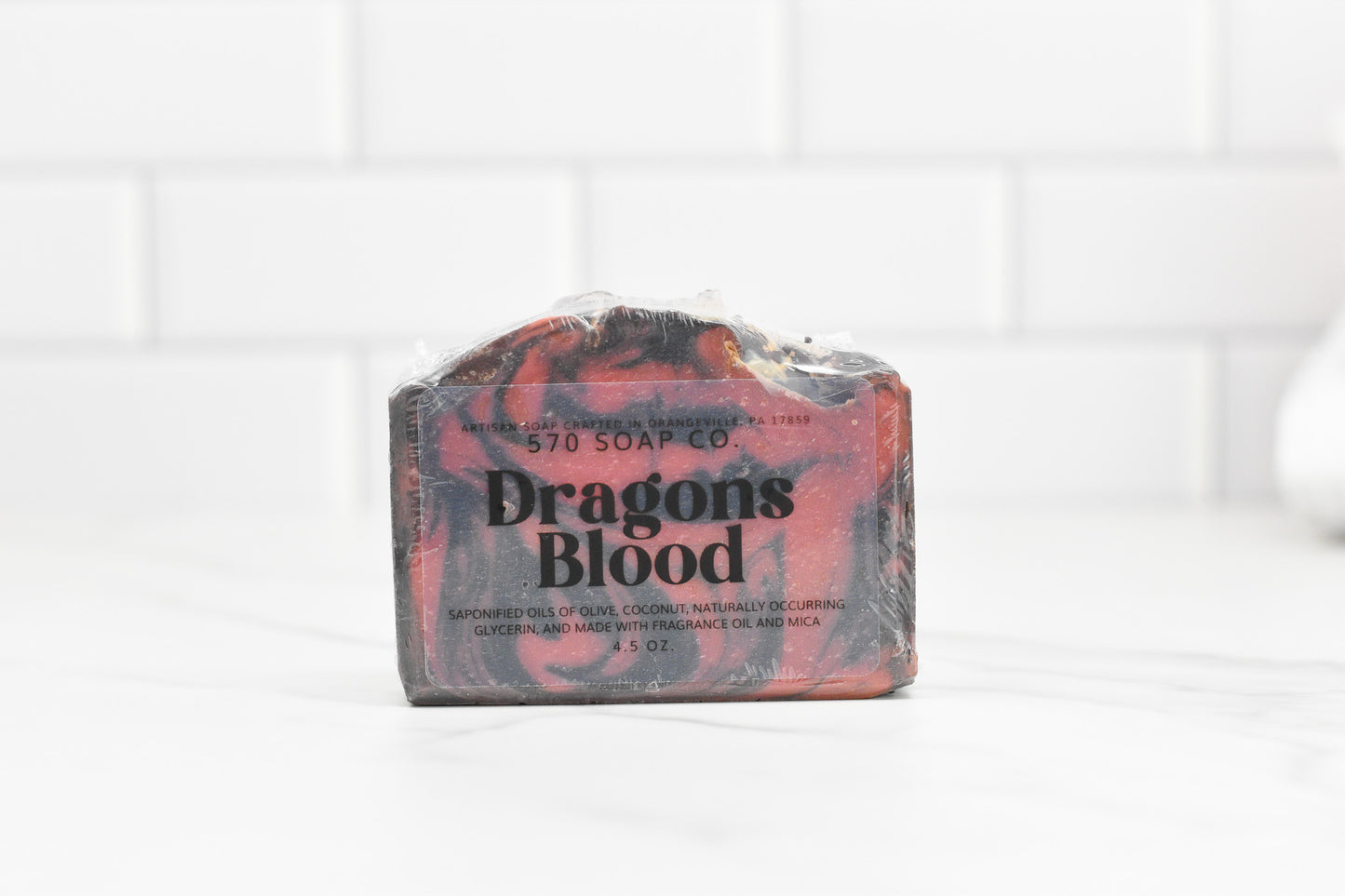 A bar of Dragons Blood Bar Soap from 570 Soap Co. features an exotic fragrance and is wrapped in plastic. Its marbled red and black hues stand out beautifully against a light-colored countertop with white tile in the background, highlighting its handcrafted quality.