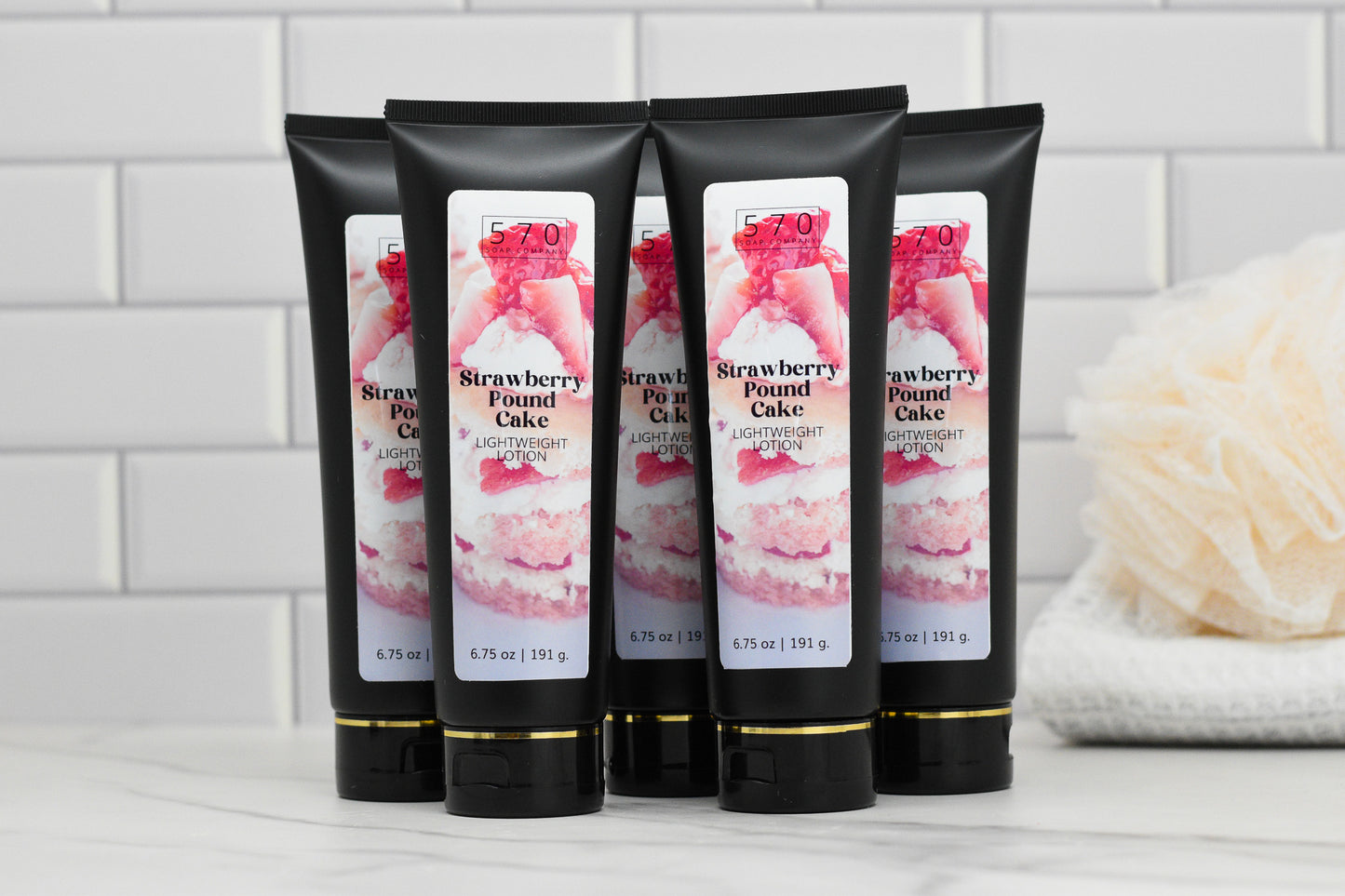 Several tubes of 570 Soap Co.'s Strawberry Pound Cake Lightweight Lotion are displayed on a bathroom counter, set against a white tiled background. A bath pouf is visible in the distance.