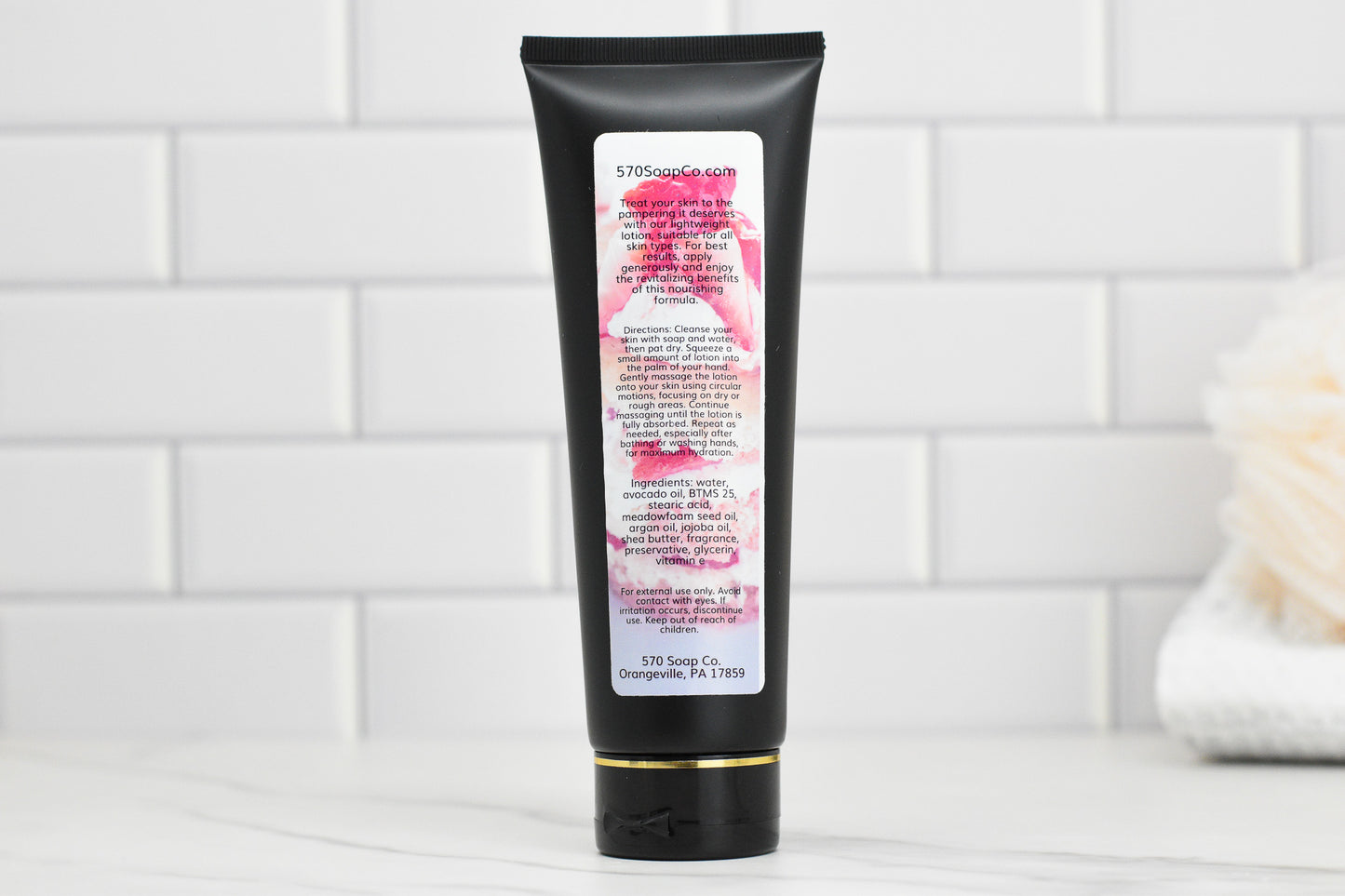 A black tube of Strawberry Pound Cake Lightweight Lotion from 570 Soap Co. stands upright on a light marble surface against a white subway tile background. The tube is adorned with text and a floral design, while a beige bath pouf can be partially seen to the right, enhancing the elegance of the scene.