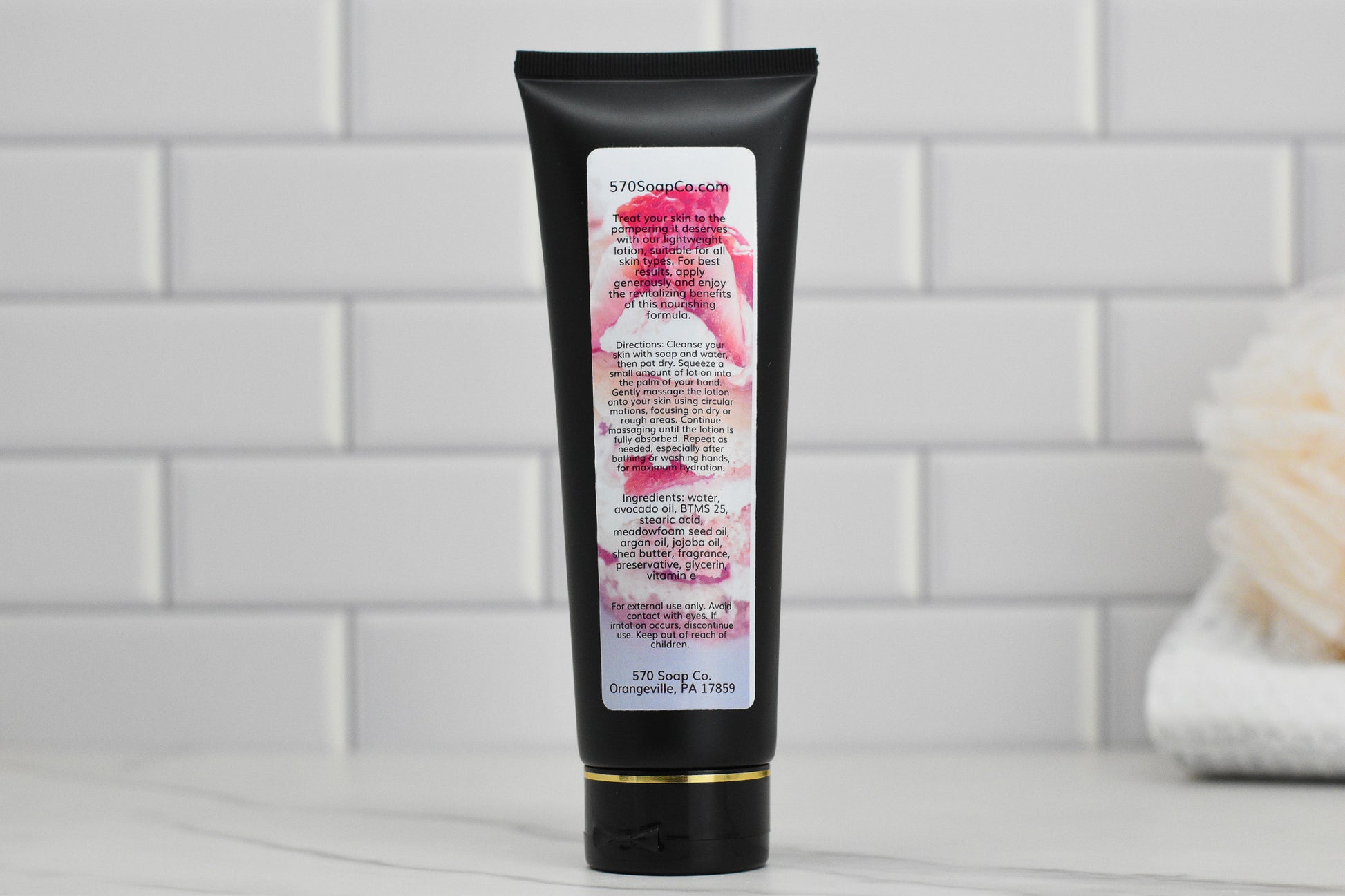 A black tube of 570 Soap Co.'s **Strawberry Pound Cake Lightweight Lotion** stands upright against a white tiled wall, exuding an aura of sweet hydration. The label features text and a floral design, while a blurred bath sponge is visible in the background on the right.