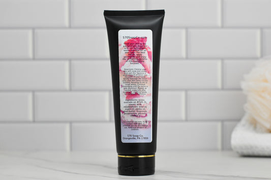 A black tube of 570 Soap Co.'s **Strawberry Pound Cake Lightweight Lotion** stands upright against a white tiled wall, exuding an aura of sweet hydration. The label features text and a floral design, while a blurred bath sponge is visible in the background on the right.
