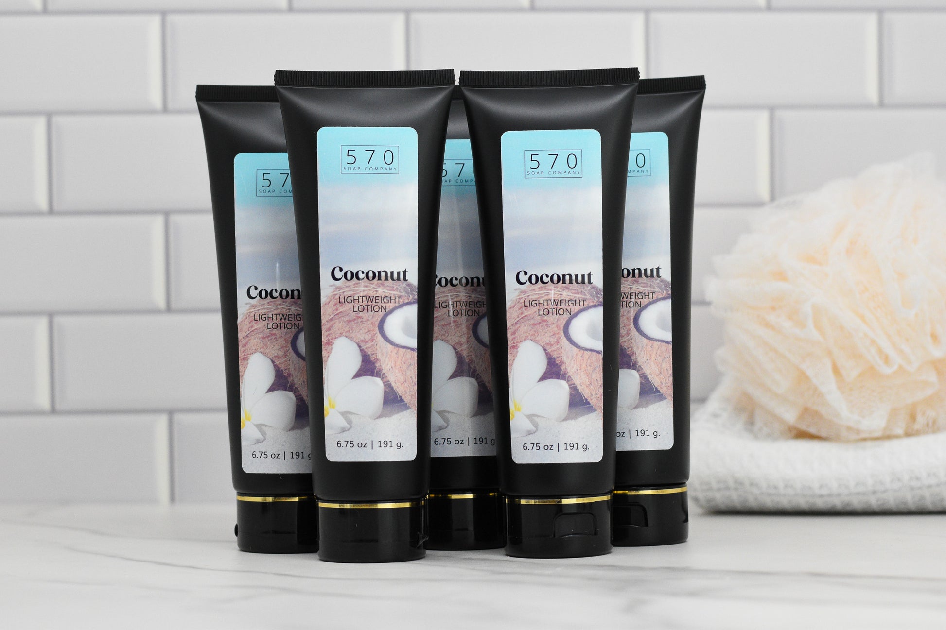 Six tubes of 570 Soap Co.'s Exotic Coconut Lightweight Lotion, providing deep hydration and a tropical paradise escape, are displayed against a white tiled background. A white bath sponge is visible in the background.