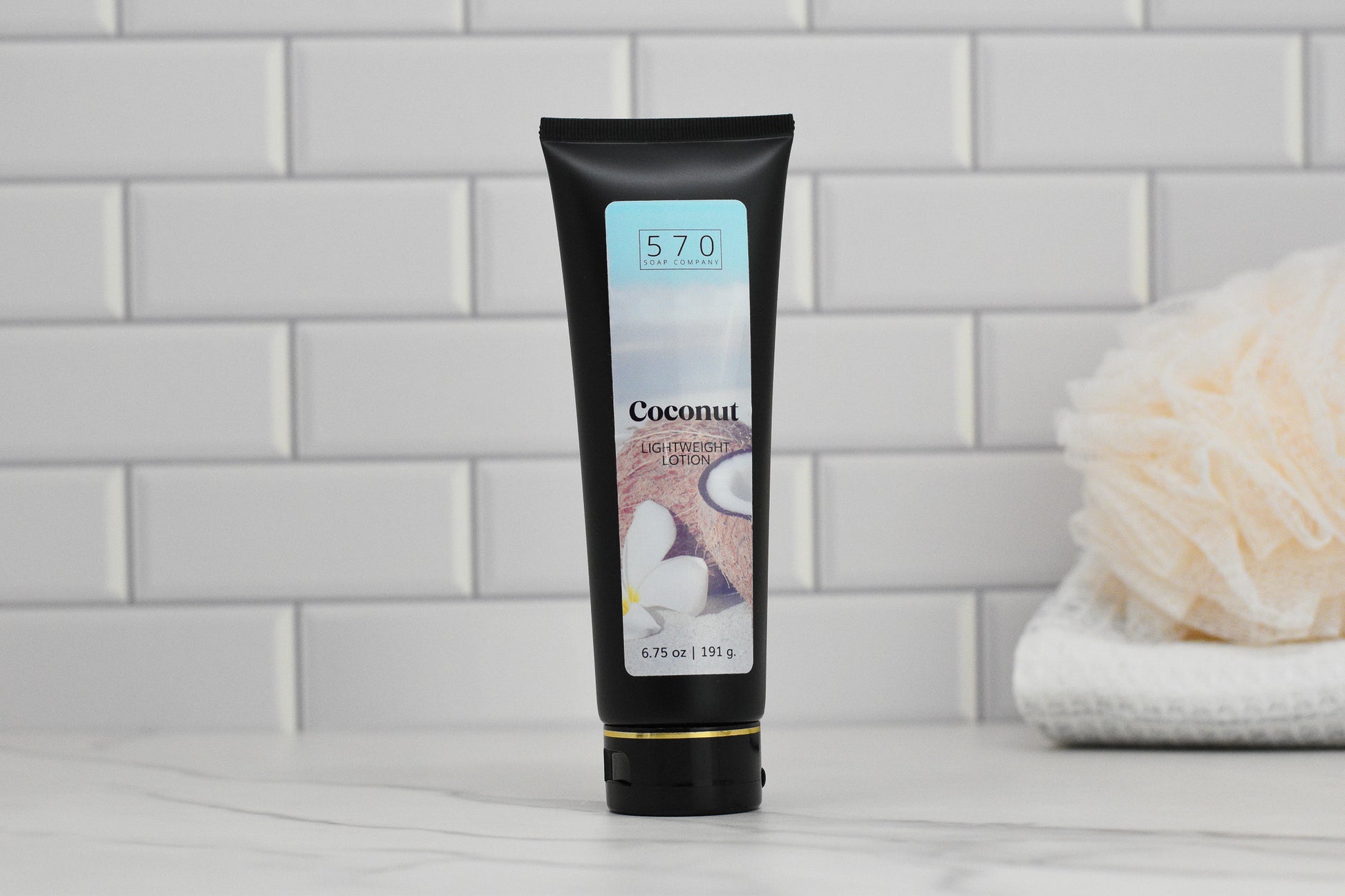 A black tube of 570 Soap Co.'s Exotic Coconut Lightweight Lotion sits on a marble countertop with its label facing outward. The background showcases a white tile wall, evoking a Tropical Paradise feel, while a beige bath pouf rests atop a stack of towels.