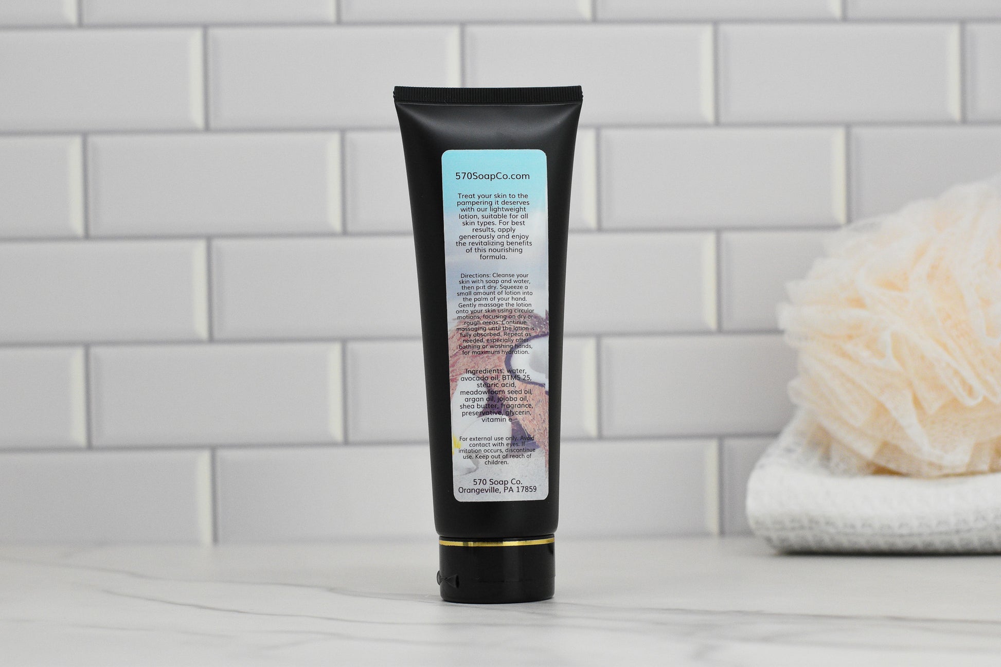 A black tube of 570 Soap Co.'s Exotic Coconut Lightweight Lotion stands upright on a tiled surface. The label is partially visible, hinting at deep hydration with tropical graphics. Nearby, a folded towel and a beige bath sponge rest on the white countertop against a backdrop of white subway tiles.