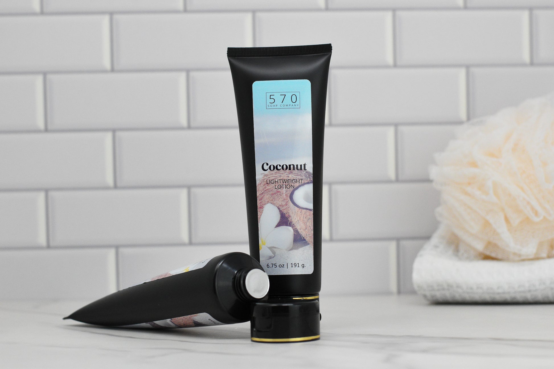 Two black tubes of Exotic Coconut Lightweight Lotion by 570 Soap Co. are on display. One stands upright, featuring a label with a coconut and flower design, while the other tube rests on its side, highlighting promises of deep hydration. In the blurred background, a white bath sponge and folded towel sit against a tiled wall, conjuring an aura of tropical paradise.