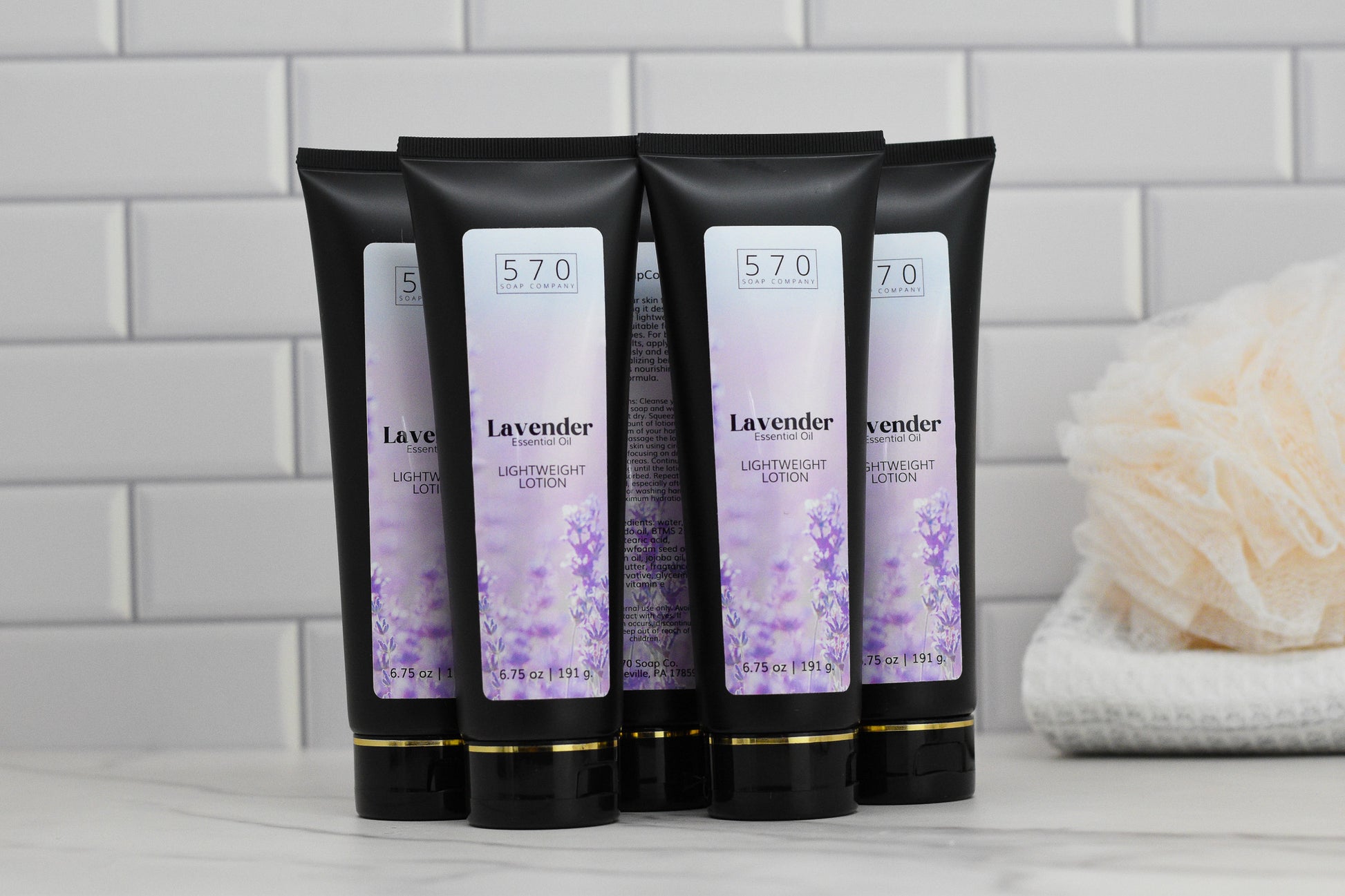 Five black tubes of 570 Soap Co.'s Lavender Lightweight Lotion - Essential Oil, featuring a calming lavender scent, are lined up in a row on a white tiled surface. In the background, a loofah and folded towel enhance the peaceful atmosphere for this moisturizing formula.