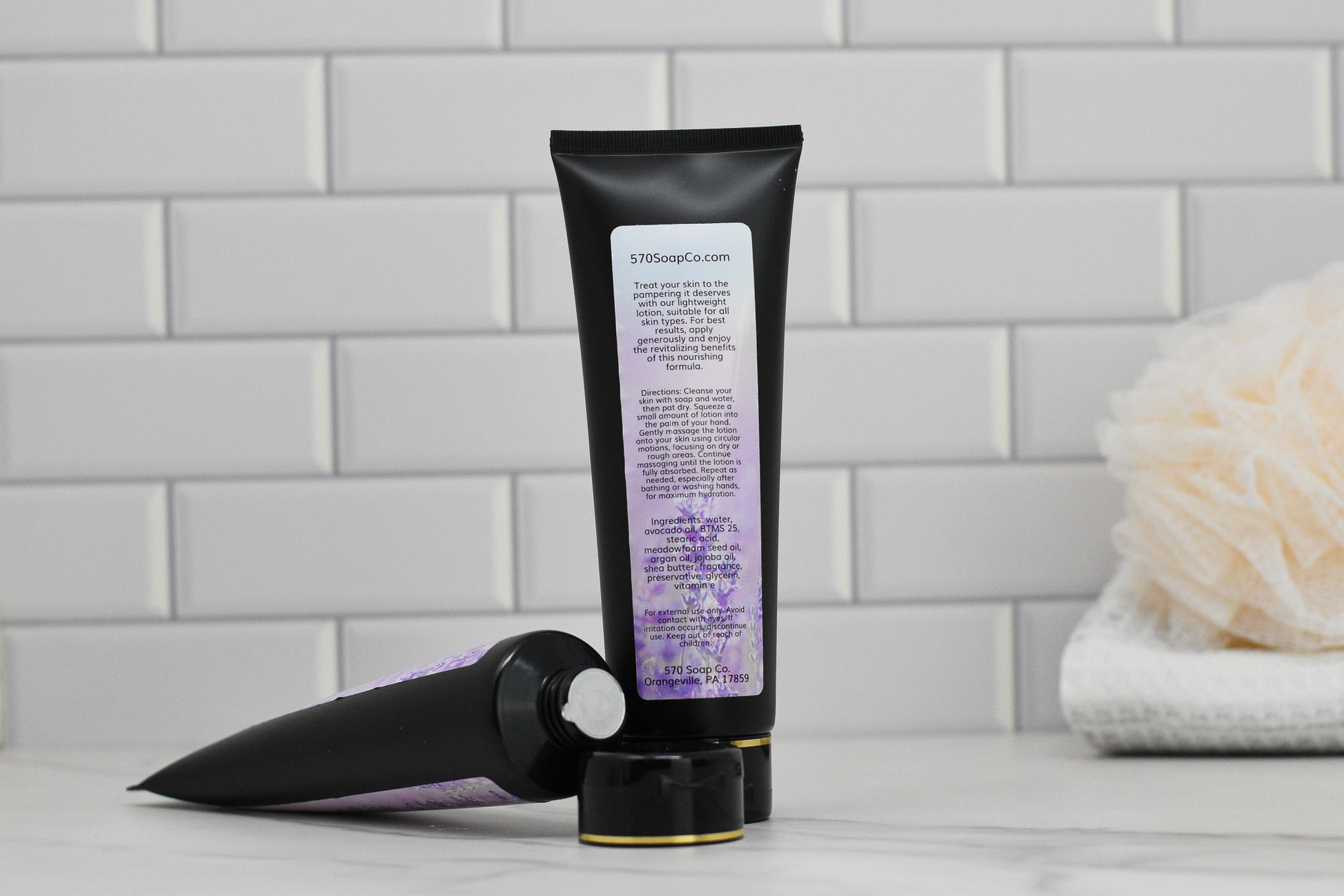 Two tubes of 570 Soap Co.'s Lavender Lightweight Lotion - Essential Oil, with distinctive purple labels and a calming lavender fragrance, are placed against a white subway tile backdrop. One tube is standing upright while the other is positioned horizontally. A bath sponge and a folded towel grace the right background, complementing the bathroom setting.