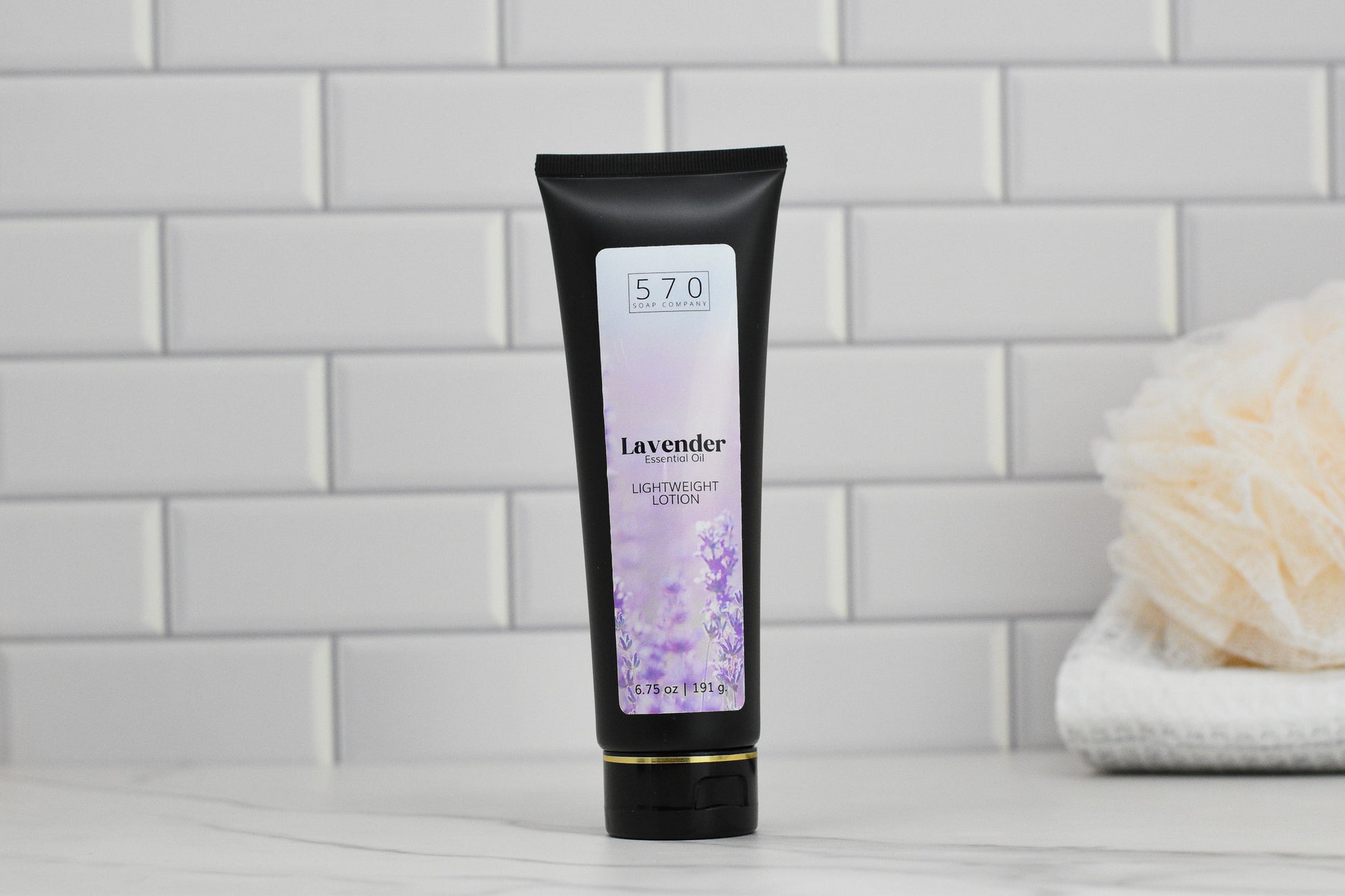 A tube of 570 Soap Co.'s Lavender Lightweight Lotion - Essential Oil, enriched with calming lavender essence, is positioned upright on a marble surface. The packaging showcases lavender flower motifs, while white tiles and a beige shower pouf beautifully enhance the tranquil setting.