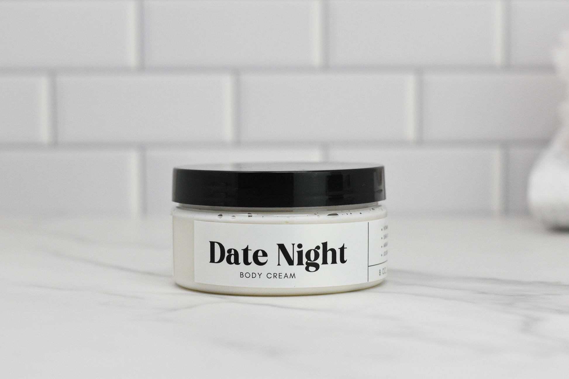 A jar of 570 Soap Co.'s Date Night Body Butter, topped with a black lid and enriched with shea butter, is elegantly placed on a marble surface against a tiled background.