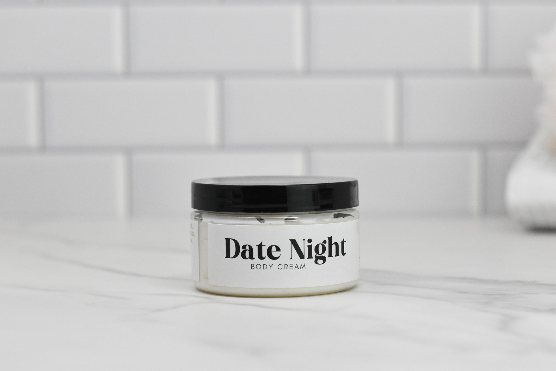 A jar of 570 Soap Co.'s Date Night Body Butter, enriched with shea butter, sits elegantly on a marble surface against a white tiled backdrop.