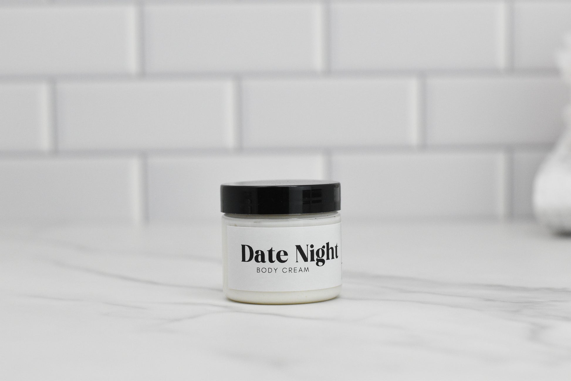 A jar of Date Night Body Butter from 570 Soap Co., infused with shea butter, rests on a marble surface, topped by a sleek black lid. The background features white subway tiles.