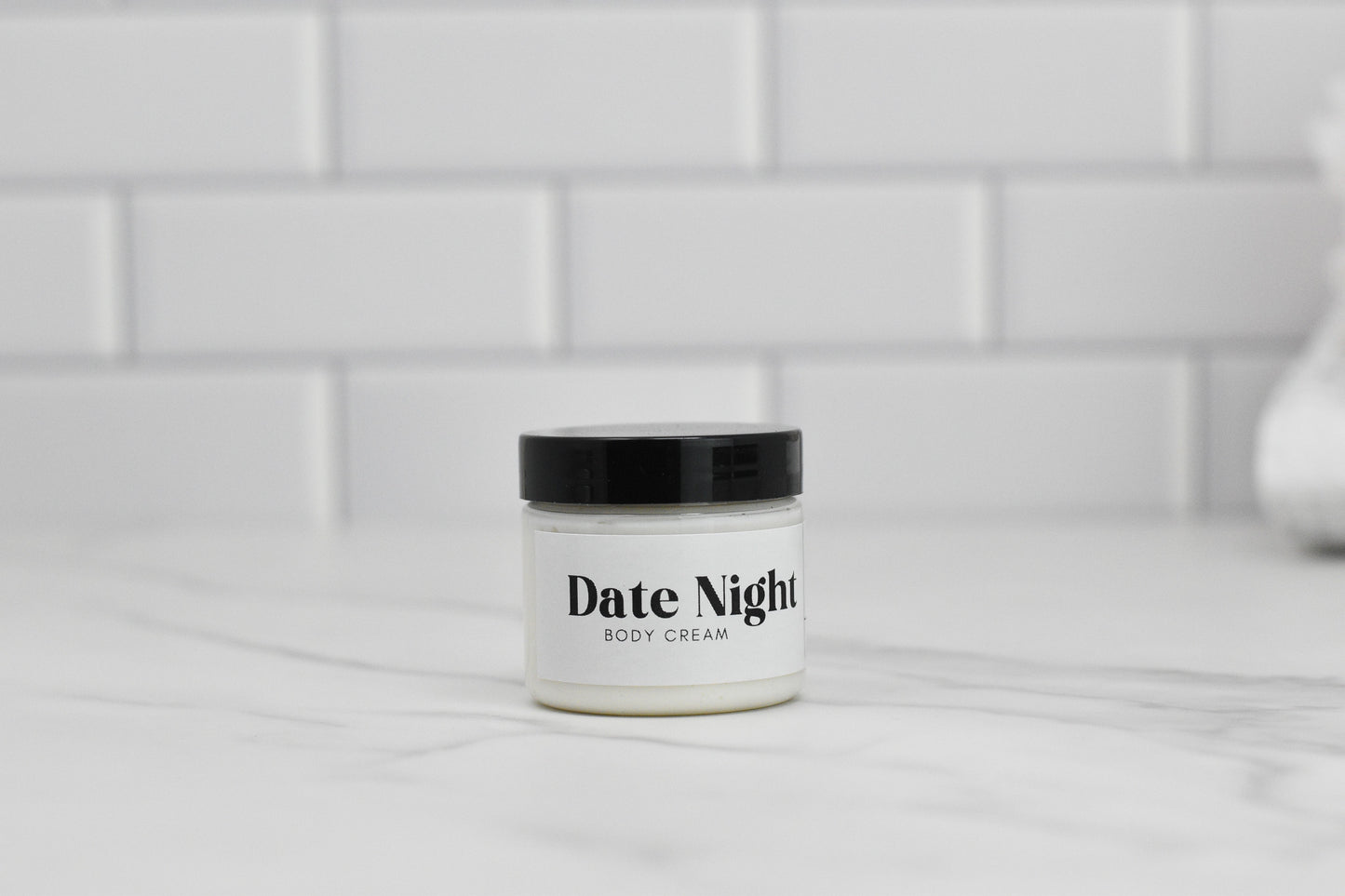 A jar of Date Night Body Butter from 570 Soap Co., infused with shea butter, rests on a marble surface, topped by a sleek black lid. The background features white subway tiles.