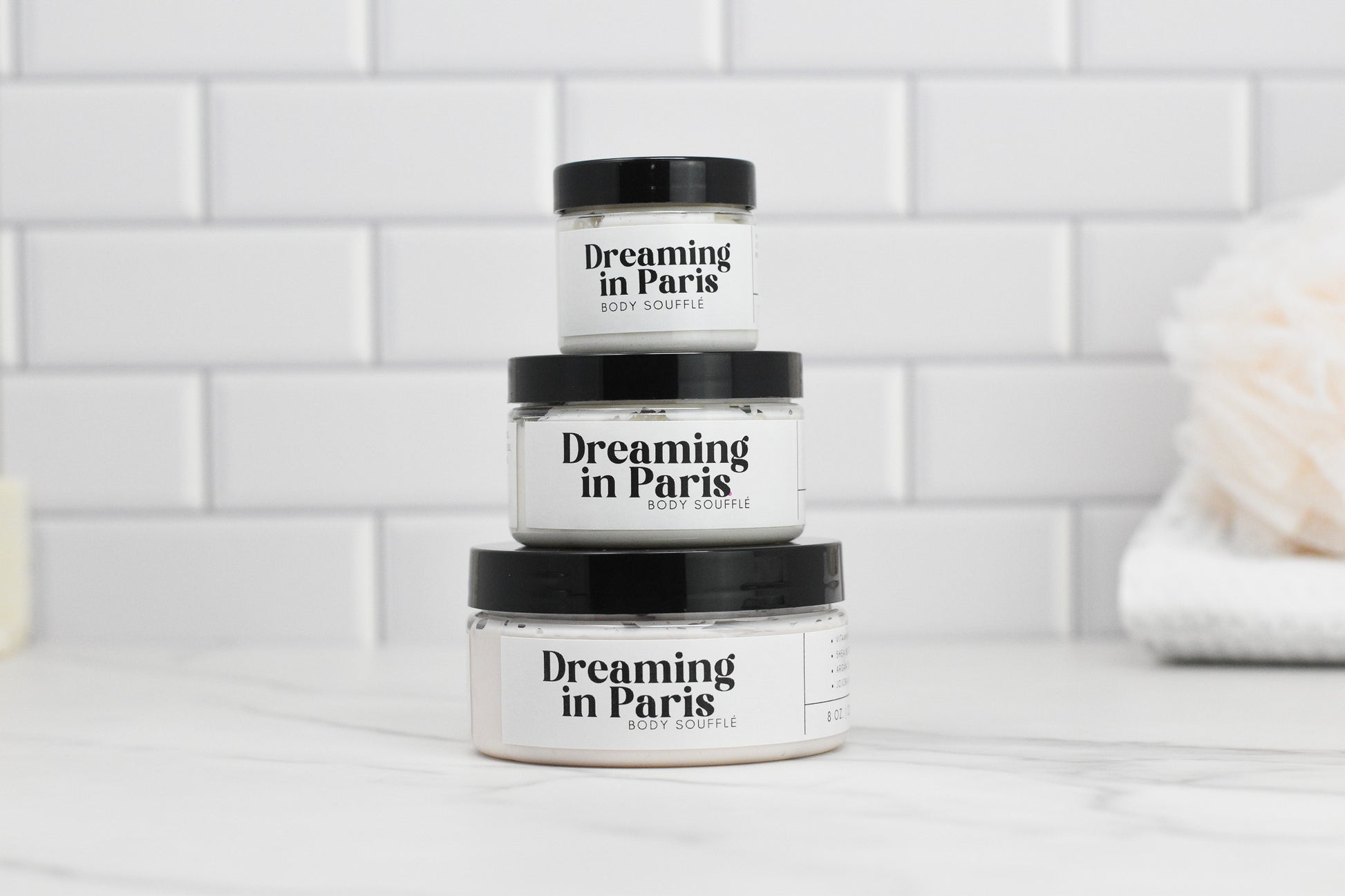 Three jars of Dreaming in Paris Body Butter by 570 Soap Co., enriched with shea butter and avocado oil, are stacked on a marble surface. The background features white subway tiles, with a soft-focus towel on the right.