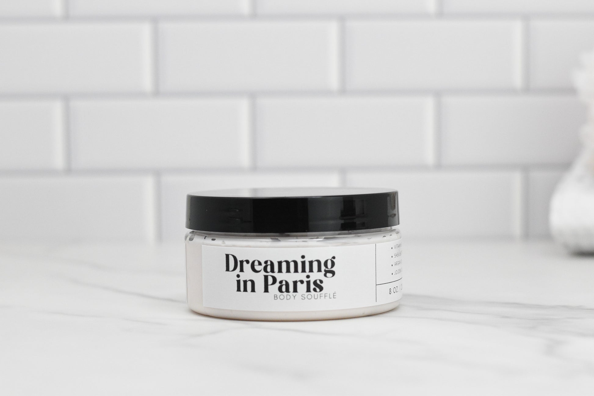 A jar of Dreaming in Paris Body Butter by 570 Soap Co., enriched with nourishing shea butter, sits elegantly on a marble surface against a white subway tile background. The jar features a sleek black lid and a label highlighting its luxurious ingredients.