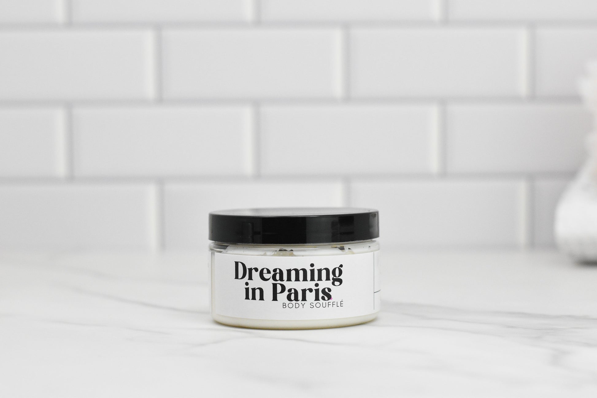 A jar of Dreaming in Paris Body Butter from 570 Soap Co., enriched with nourishing shea butter, is elegantly placed on a white marble surface. The background features a white tiled wall, creating a clean and minimalistic aesthetic.