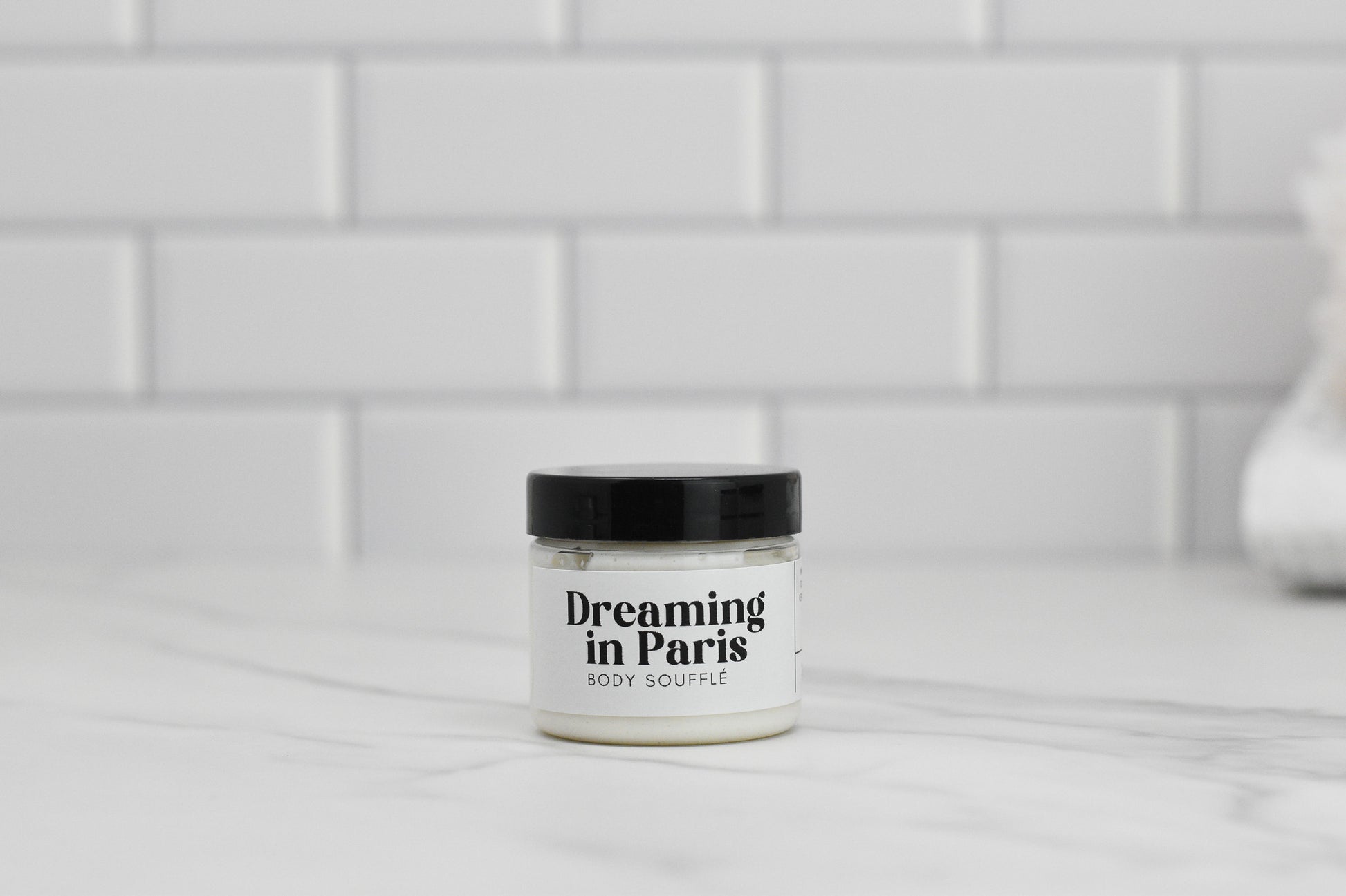 A jar of 570 Soap Co.'s Dreaming in Paris Body Butter, enriched with avocado oil, rests elegantly on a white marble surface. The sleek container features a black lid and stands before a pristine white tiled backsplash, promising a luxurious skincare experience.