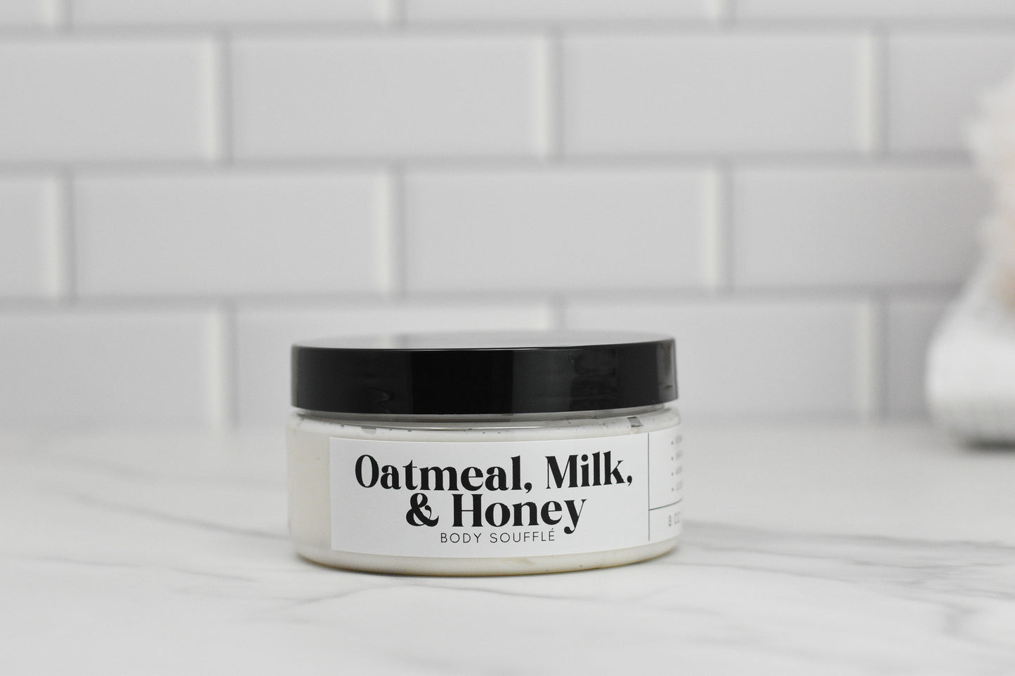 A jar of Oatmeal Milk & Honey Body Butter by 570 Soap Co., featuring a black lid, is elegantly positioned on a white marble surface. Enriched with nourishing shea butter, it embodies luxury. Behind it, a white tiled wall enhances the clean and minimalist aesthetic.