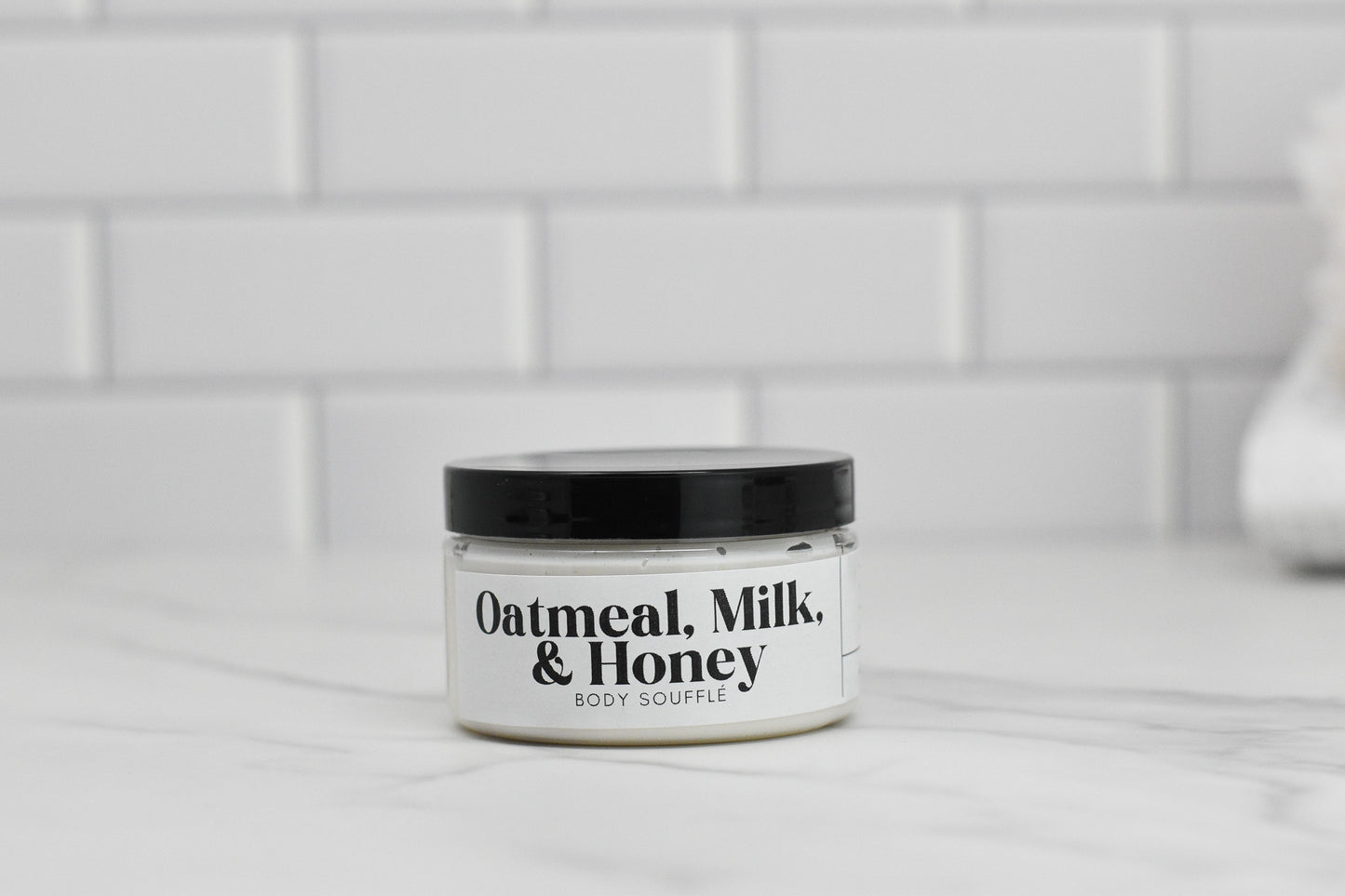 A jar of 570 Soap Co.'s Oatmeal Milk & Honey Body Butter, adorned with a black lid, sits elegantly on a white marble surface. Enriched with luxurious shea butter, the background is enhanced by sleek white subway tiles.