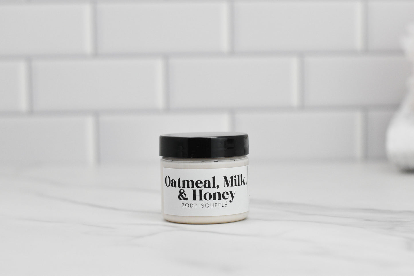 On a marble countertop against a backdrop of white subway tiles, a jar of Oatmeal Milk & Honey Body Butter from 570 Soap Co. is displayed. It features a black lid and a minimalist label design, promising luxurious hydration.
