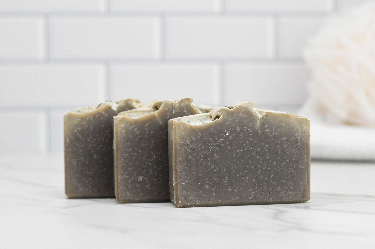 Three bars of 570 Soap Co.'s Lavender Essential Oil Bar Soap - All Natural rest on a marble surface against a white tile backdrop. These handmade soaps feature a speckled texture with an uneven top edge. A fluffy bath sponge is partially visible in the background.