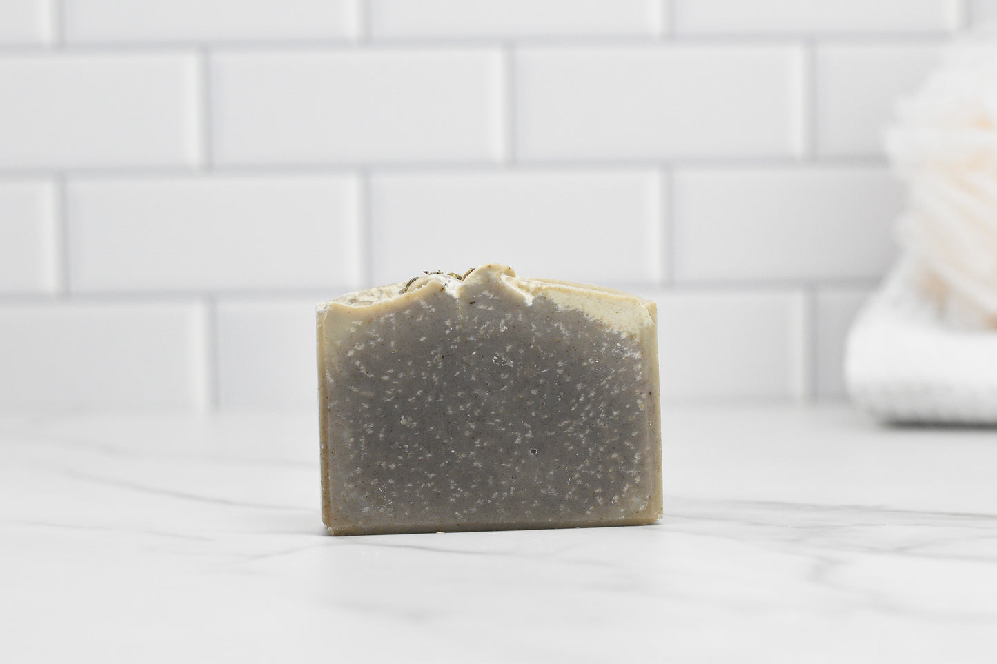 A bar of Lavender Essential Oil Bar Soap - All Natural by 570 Soap Co., featuring a speckled gray hue from infused kaolin clay and an uneven top, rests elegantly on a marble surface. The background showcases a white subway tile wall, enhanced by the soothing presence of lavender essential oil, creating a clean, minimalistic aesthetic.