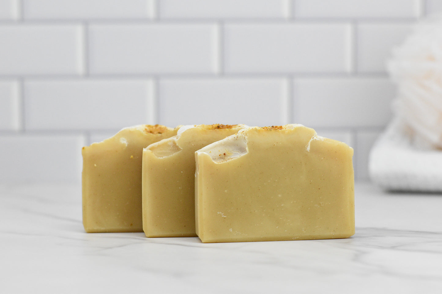 Three bars of the Orange Patchouli Bar Soap - All Natural by 570 Soap Co., with their light beige hue and textured tops, are placed on a white marble surface. The backdrop consists of a white tiled wall, highlighting the clean and minimalist style.