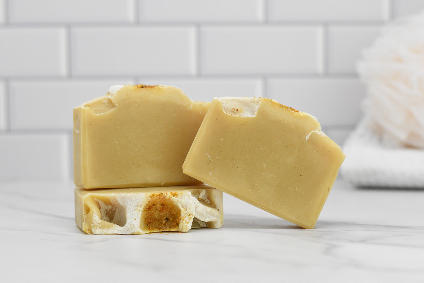 Three bars of 570 Soap Co.'s Orange Patchouli Bar Soap - All Natural are stacked on a white marble surface in front of a white tiled background. The light beige soap, featuring subtle notes of Orange Patchouli Essential Oil, has a speckled appearance and a textured top, with a bath sponge visible in the background.