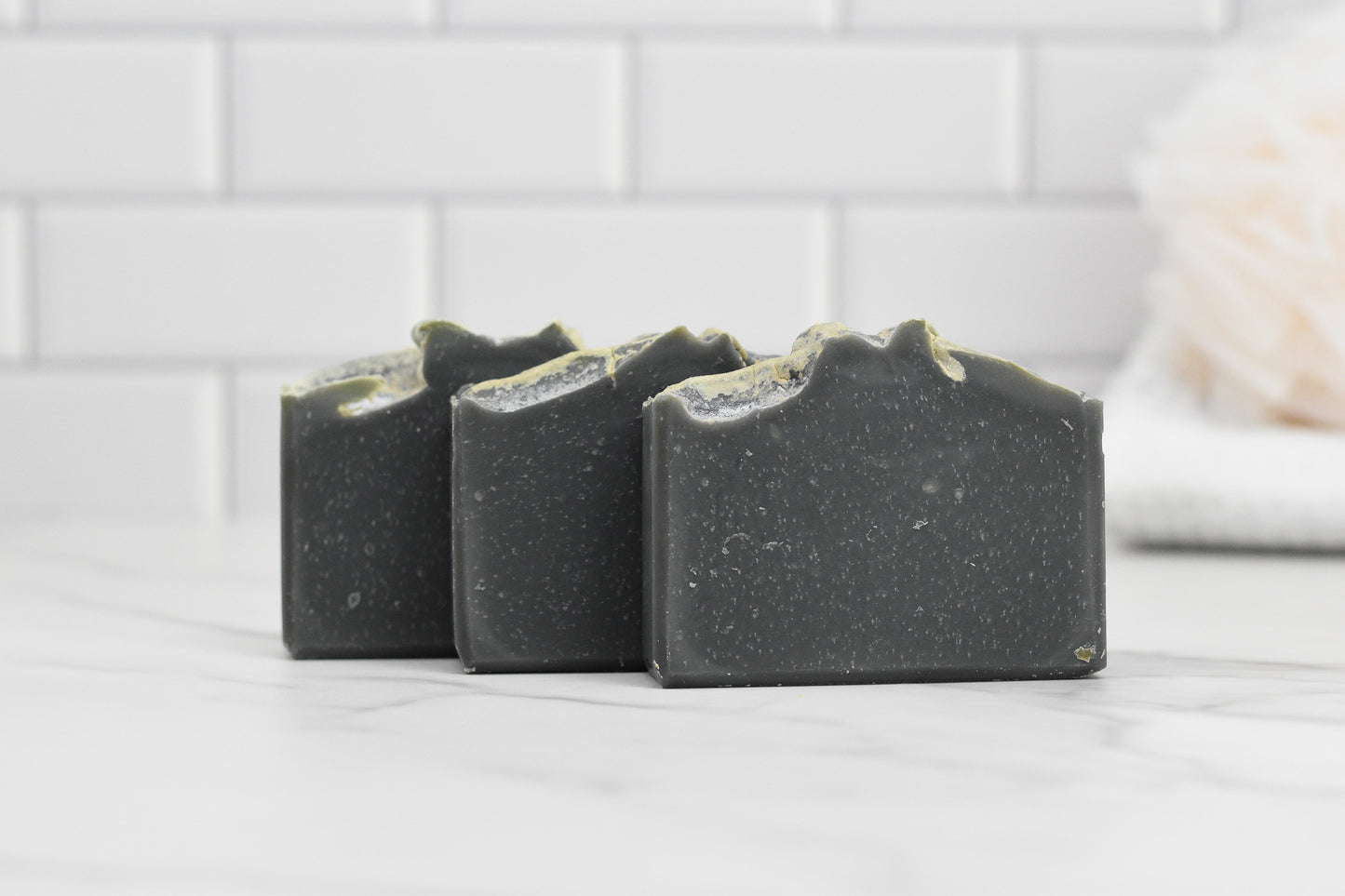 Three bars of 570 Soap Co.'s Kentucky Bourbon Charcoal Bar Soap, each in a dark gray hue with textured tops, are displayed on a white marble surface. The background showcases a crisp white subway tile wall, offering Southern elegance with its clean and minimalist aesthetic.