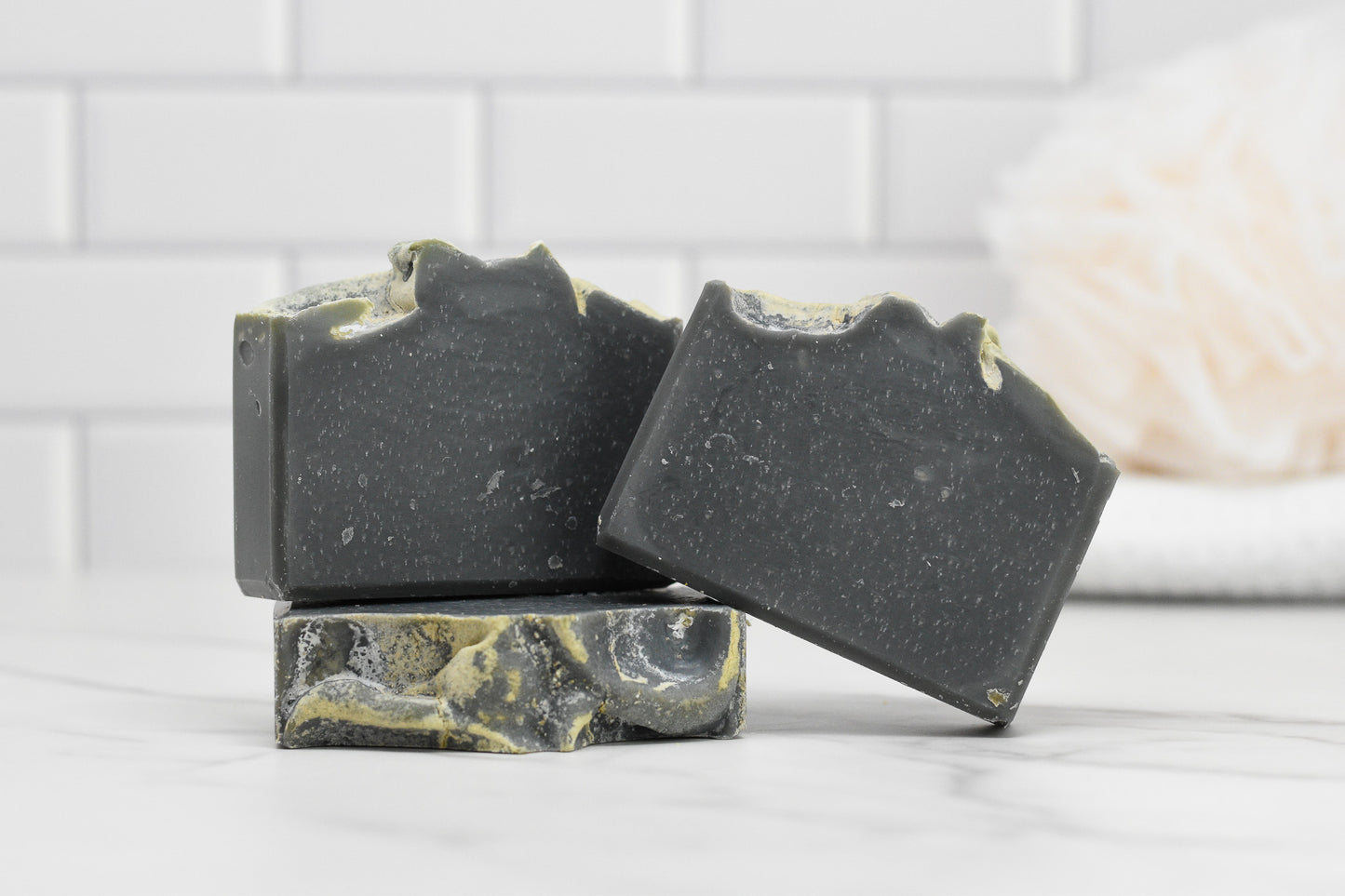 Three bars of the Kentucky Bourbon Charcoal Bar Soap from 570 Soap Co. are stacked creatively on a white marble surface. The background features white subway tiles and a blurred soft sponge, enhancing the clean, minimalist aesthetic while adding a touch of Southern elegance.
