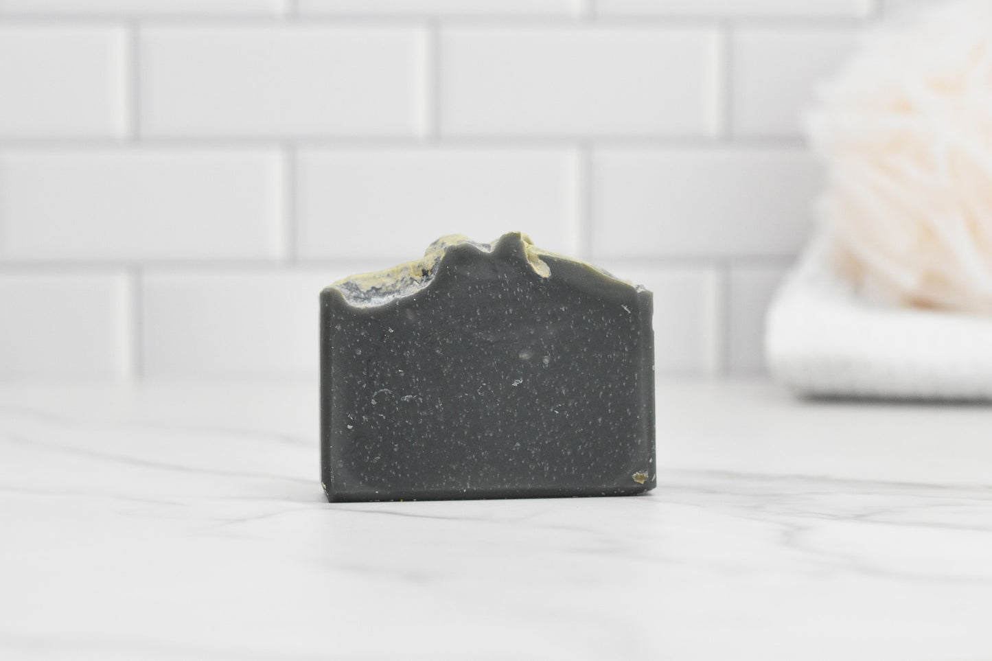 A black bar of Kentucky Bourbon Charcoal Bar Soap by 570 Soap Co. with textured edges rests on a white marble countertop. The background showcases white subway tiles and a blurred beige loofah, enhancing the scene with Southern elegance.