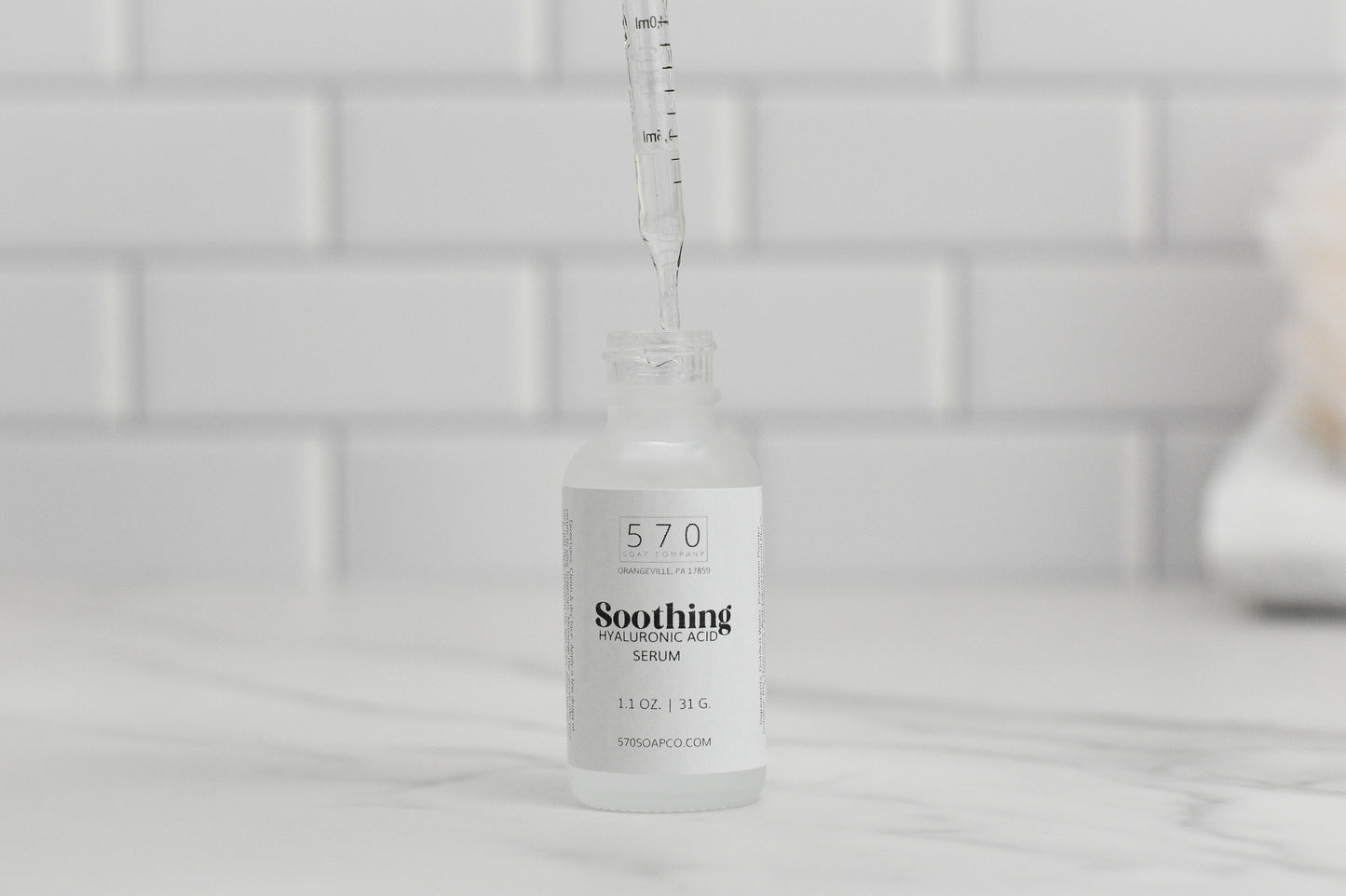 A dropper delicately dispenses liquid into a clear glass bottle labeled "Soothing Hyaluronic Acid Serum by 570 Soap Co." on a marble surface. The backdrop of white subway tiles enhances the sense of purity as the serum hydrates and soothes your skin.
