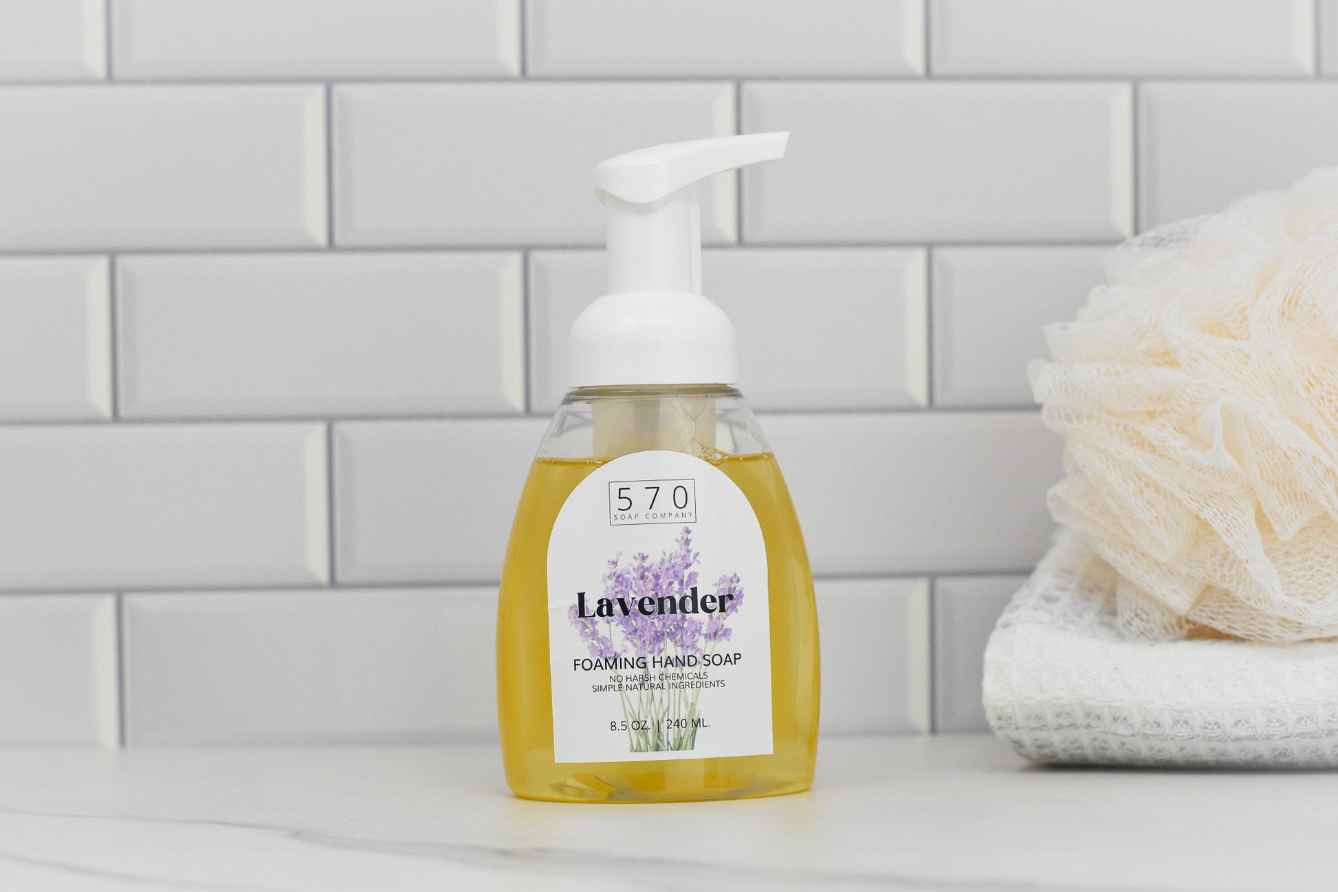 A bottle of 570 Soap Co. Lavender Natural Foaming Hand Soap - Made with Essential Oils sits on a white countertop, against a backdrop of white subway tiles. A beige loofah and folded towels are to the right.
