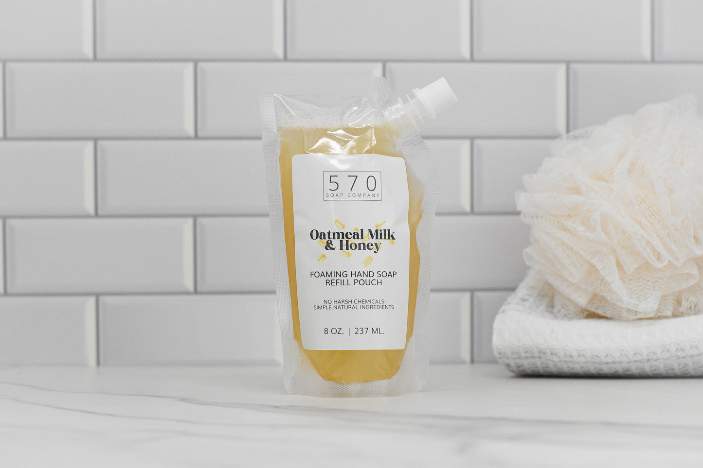 A refill pouch of Oatmeal Milk & Honey Scented Foaming Hand Soap from 570 Soap Co. is displayed on a white countertop, providing moisturizing benefits. In the background, a white loofah and folded towel rest against a tiled wall.