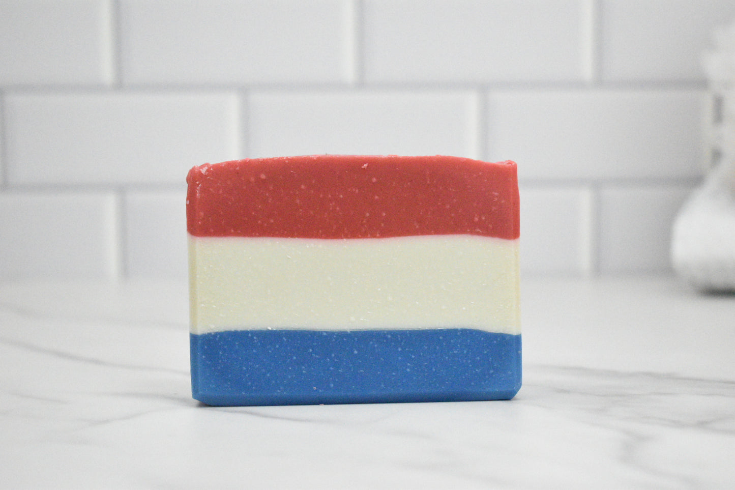 Three-layered soap bar with red, white, and blue stripes on a white tiled background