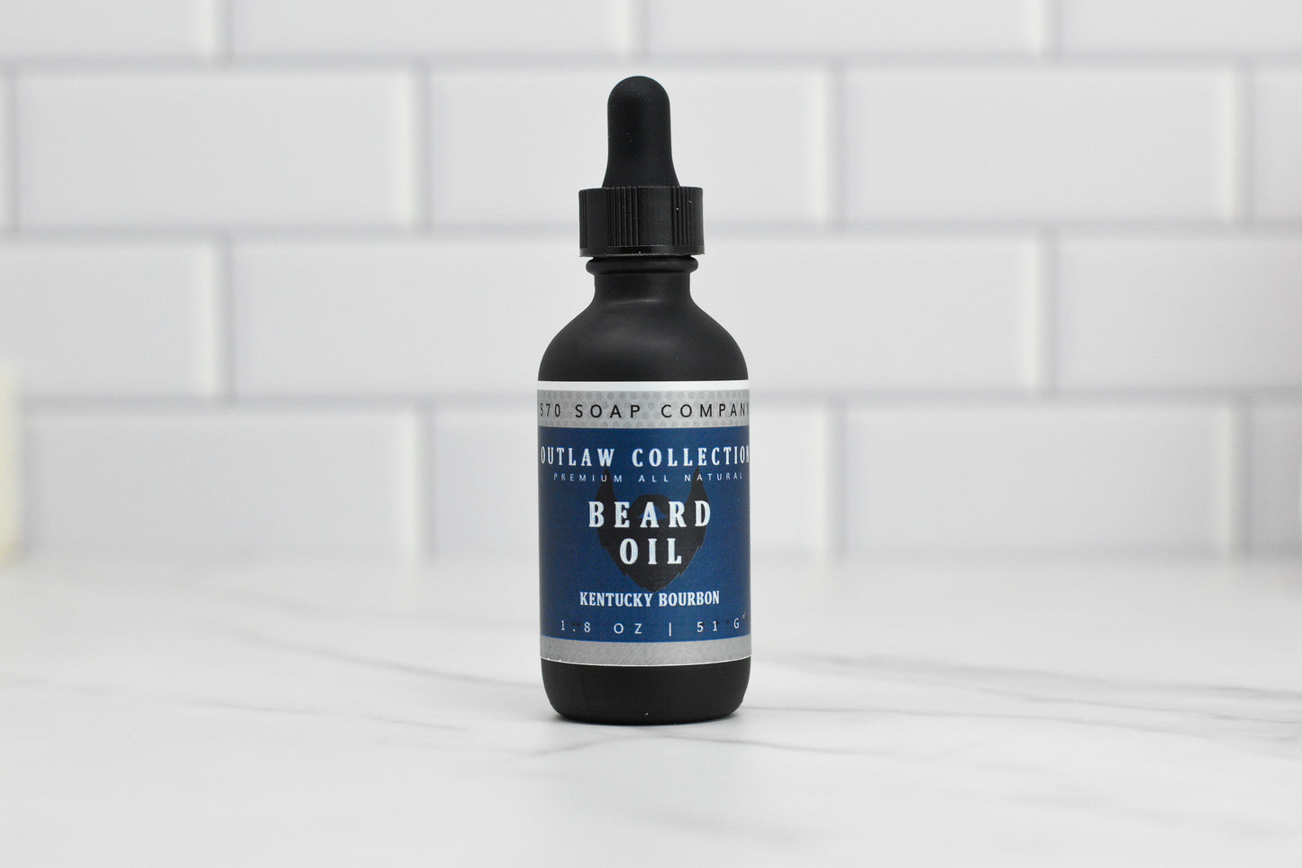A bottle of 570 Soap Co.'s Beard Oil - Choose your scent, featuring a black dropper cap and labeled as part of the Outlaw Collection with a Kentucky Bourbon fragrance, is crafted using natural oils. Placed elegantly on a white marble surface with a blurred tile background, it adds the perfect touch to your beard care routine.