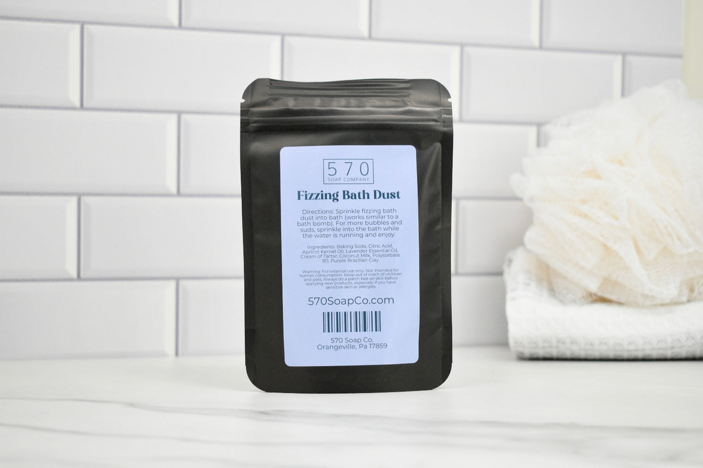 Black packaging of 570 Fizzing Bath Dust with a white label on a white tiled background.