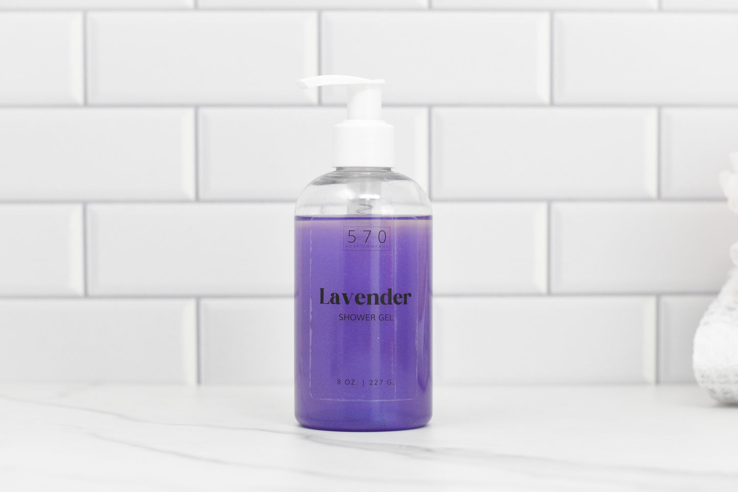 A clear pump bottle filled with luxurious Bubble Bliss Lavender Shower Gel from 570 Soap Co.'s exclusive collection rests on a white marble surface, with the backdrop of white subway tiles enhancing the clean, minimalistic aesthetic of this premium bath essential.