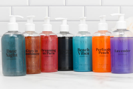 A lineup of seven Bubble Bliss Luxury Shower Gels from 570 Soap Co. adorns a white countertop, each bottle showcasing a unique color and label from an enticing fragrance collection: Date Night, Cozy in Cashmere, Dreaming in Paris, Midnight Amber, Beach Vibes, Perfectly Peach, and Lavender. A tiled wall provides the backdrop.
