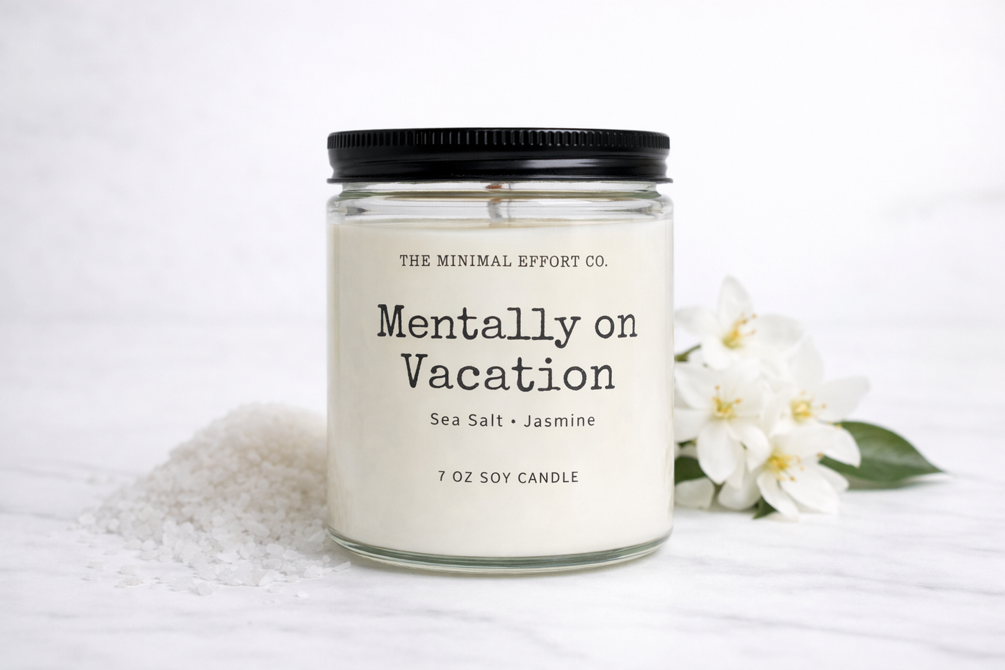 Candle labeled 'Mentally on Vacation' from The Minimal Effort Co. with a white flower and towel in the background.