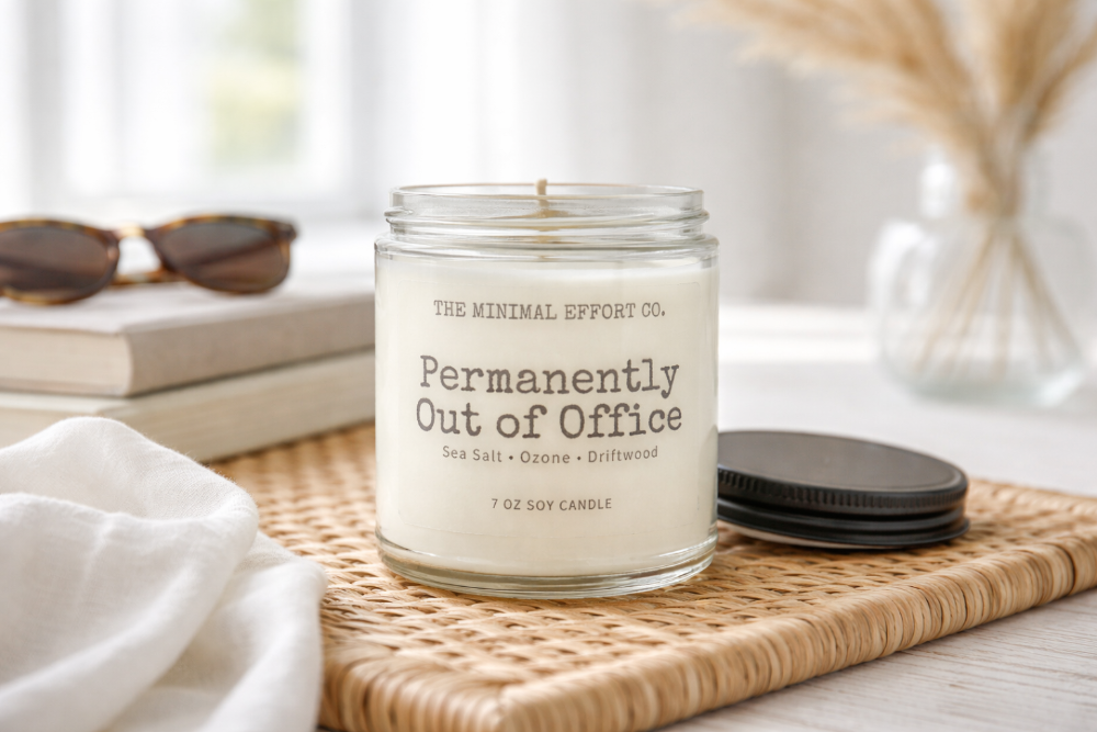 Candle labeled 'Permanently Out of Office' by The Minimal Effort Co. on a woven mat with sunglasses and books in the background.