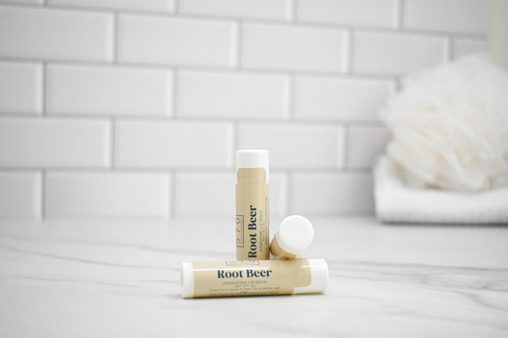 Two lip balm sticks labeled 'Root Beer' on a white surface with a tiled wall background.