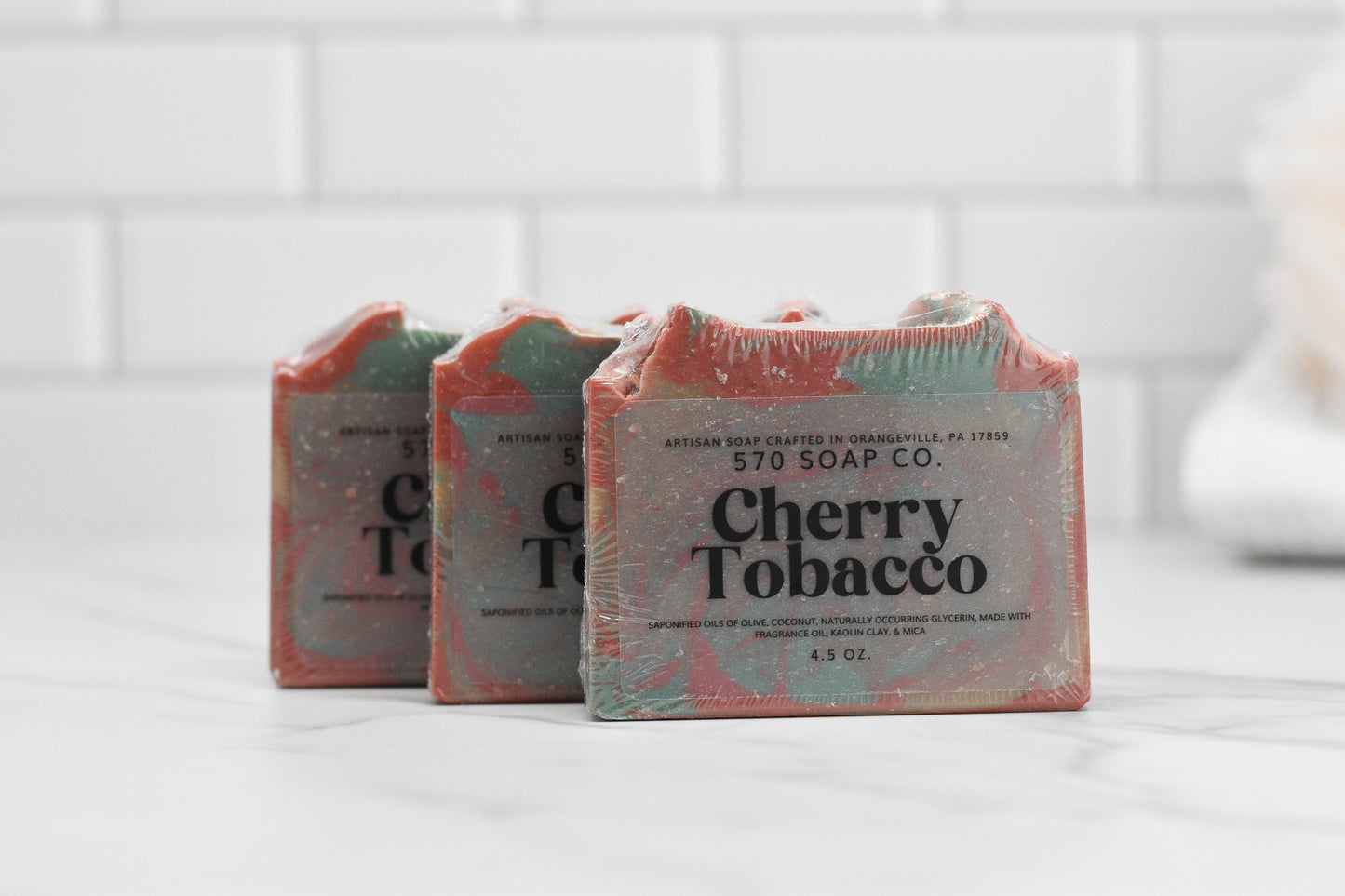 A trio of Cherry Tobacco Bar Soaps from 570 Soap Co. rests elegantly on a white marble surface. Their rustic packaging, showcasing text about the soap's crafting location, beautifully contrasts with the light tiled background. The inclusion of kaolin clay adds to its artisanal allure.