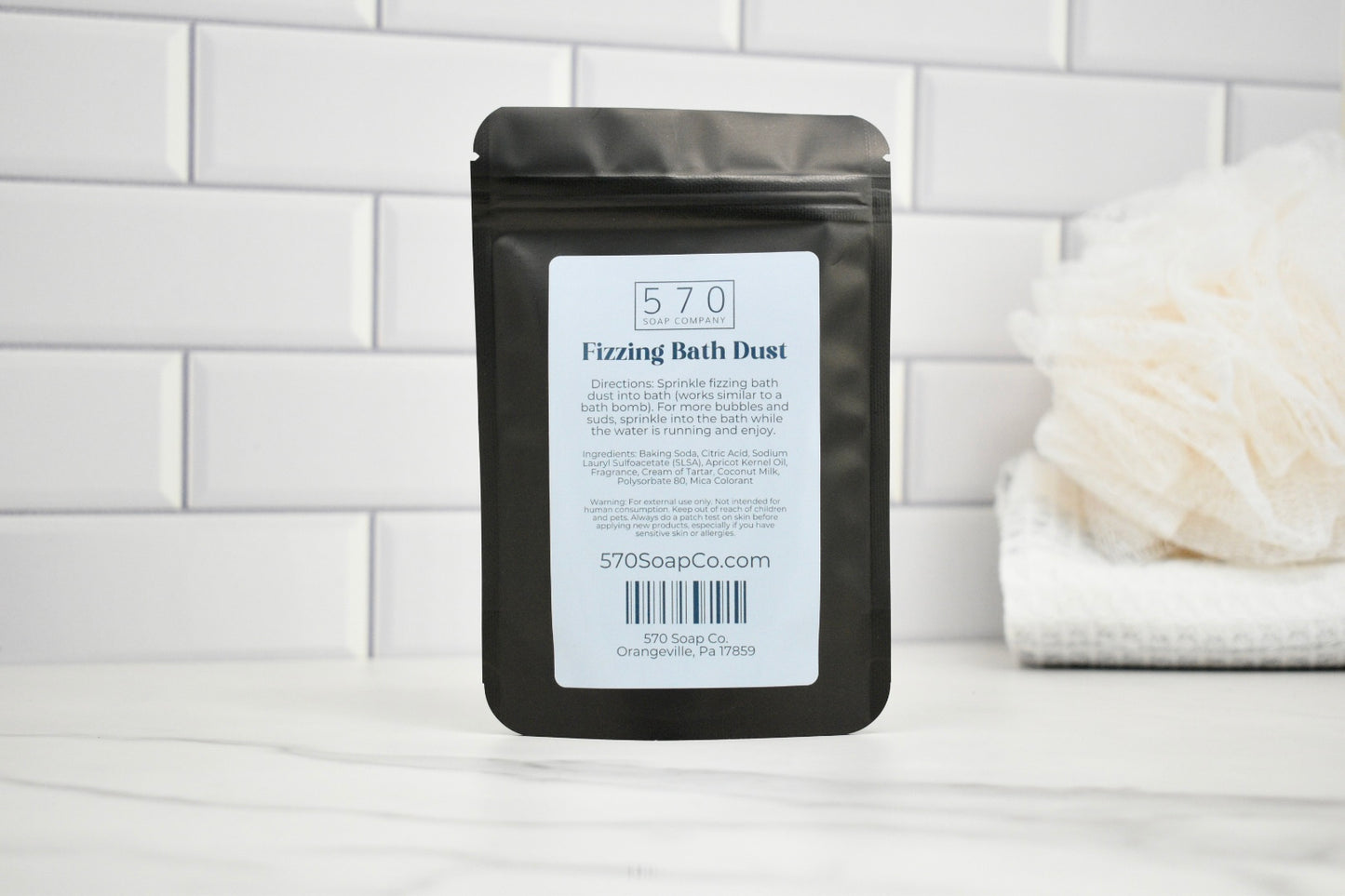 Packaging of 570 Fizzing Bath Dust on a bathroom counter with a white towel and bath bomb in the background.