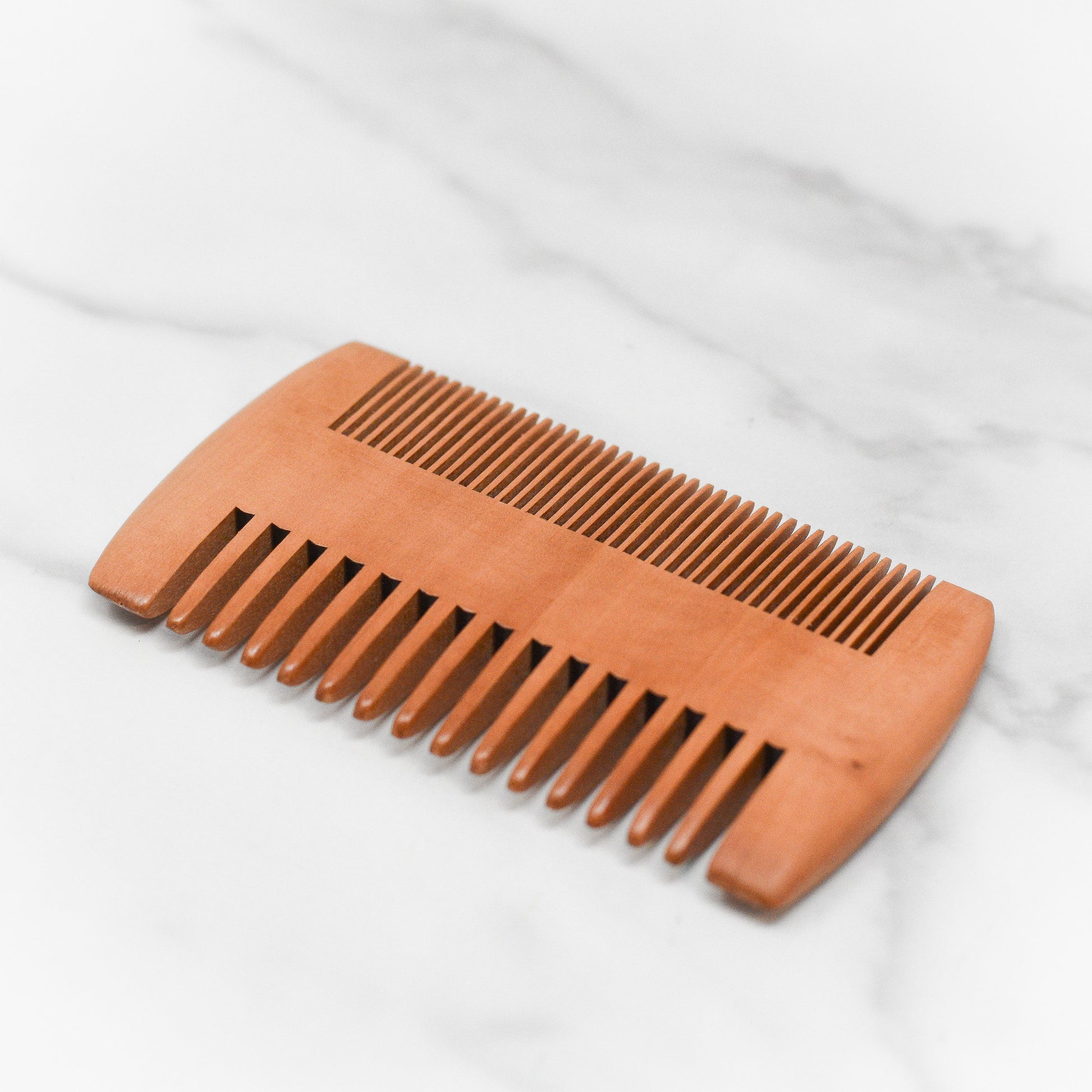 Introducing the Beard Grooming Tools from 570 Soap Co.—a dual-sided wooden comb featuring fine teeth on one side and wide teeth on the other, ideal for grooming. Elegantly displayed on a smooth marble surface, it embodies an essential in beard care essentials.