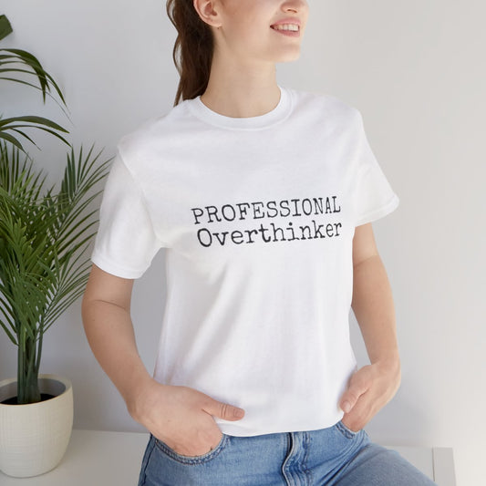 Person wearing a white t-shirt with 'PROFESSIONAL Overthinker' text, standing indoors next to a plant.