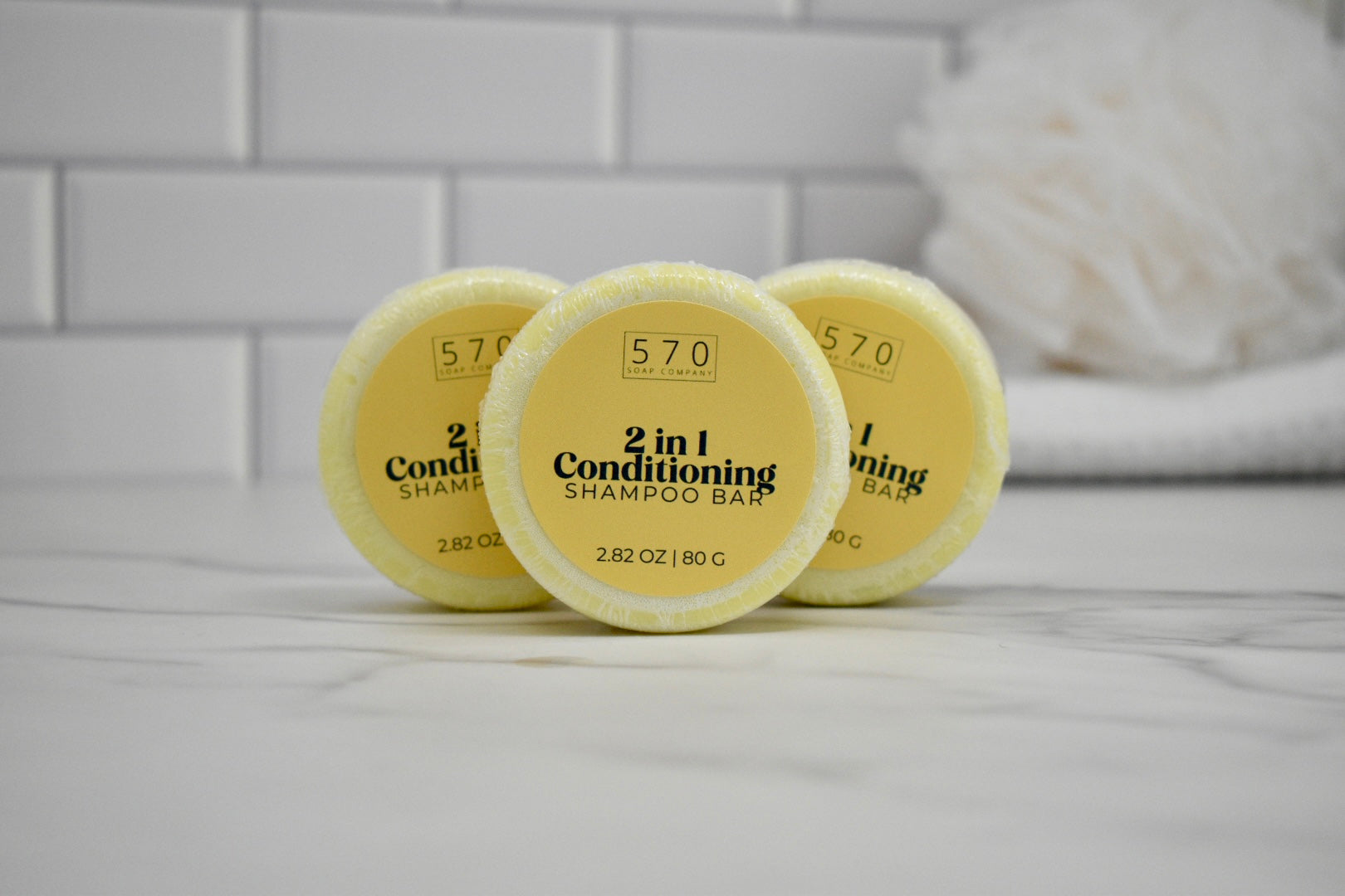Three yellow 2-in-1 conditioning shampoo bars on a white surface with a tiled wall background.