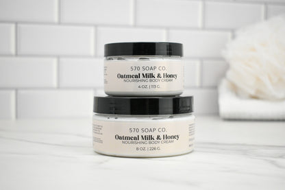 Two jars of Oatmeal Milk & Honey body cream from 570 Soap Co. on a white surface with a tiled wall background.