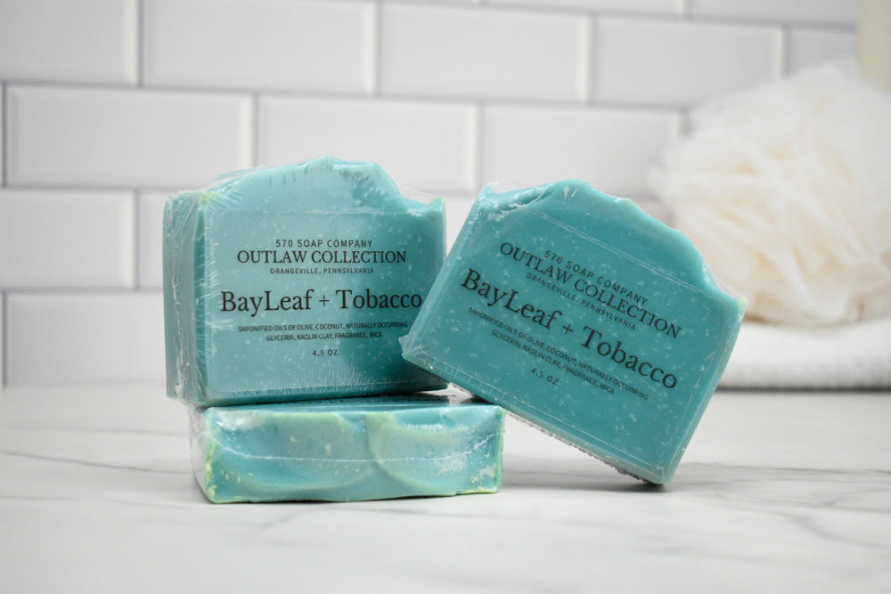 Three bars of soap labeled 'BayLeaf + Tobacco' from the Outlaw Collection on a white surface with a tiled wall background.